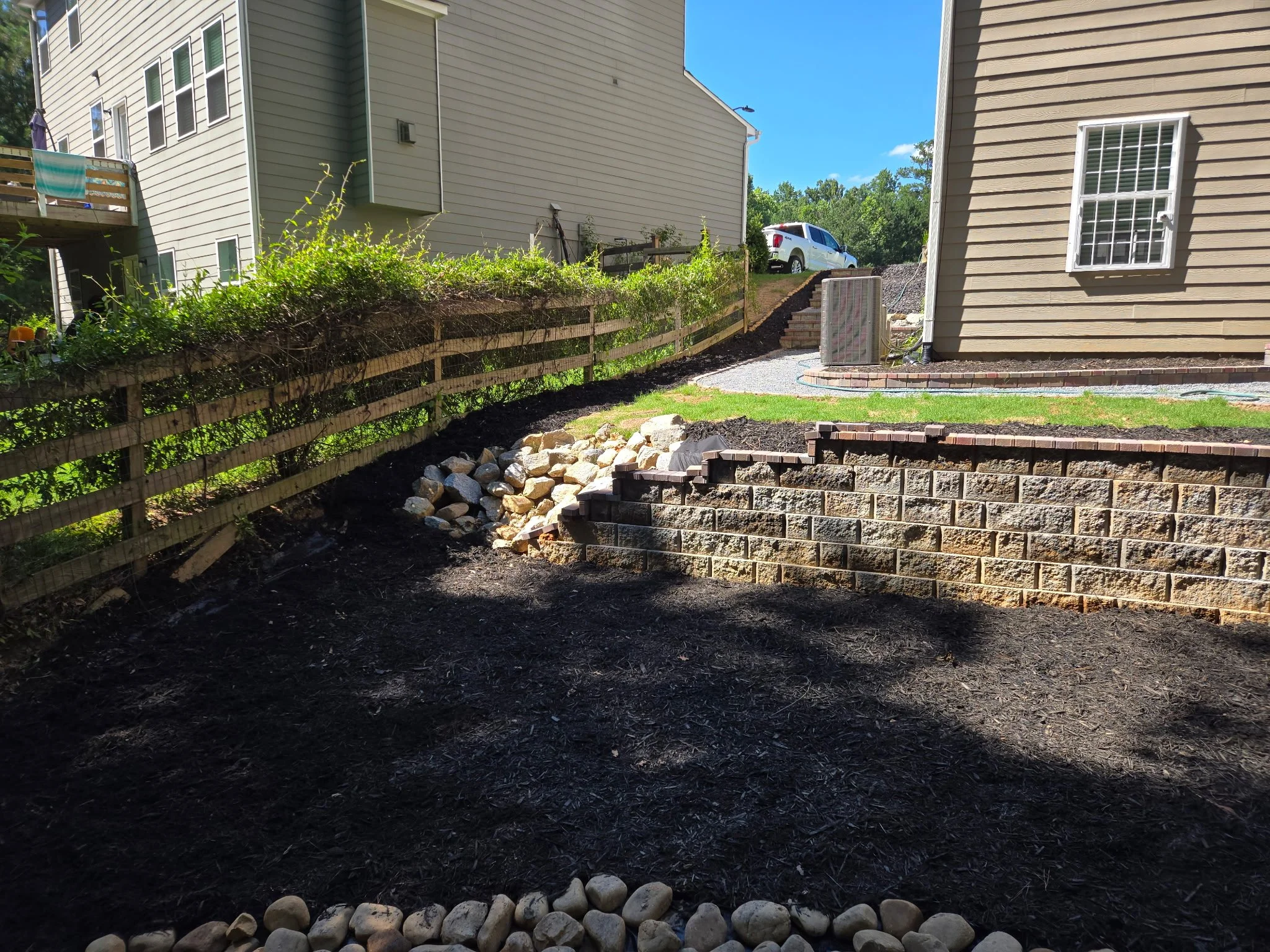 Backyard with terraced landscaping, brick retaining wall, rocks, a wooden fence, shrubbery, a house with vinyl siding, a window, and an air conditioning unit. A white truck is parked on the hill in the background under a blue sky.