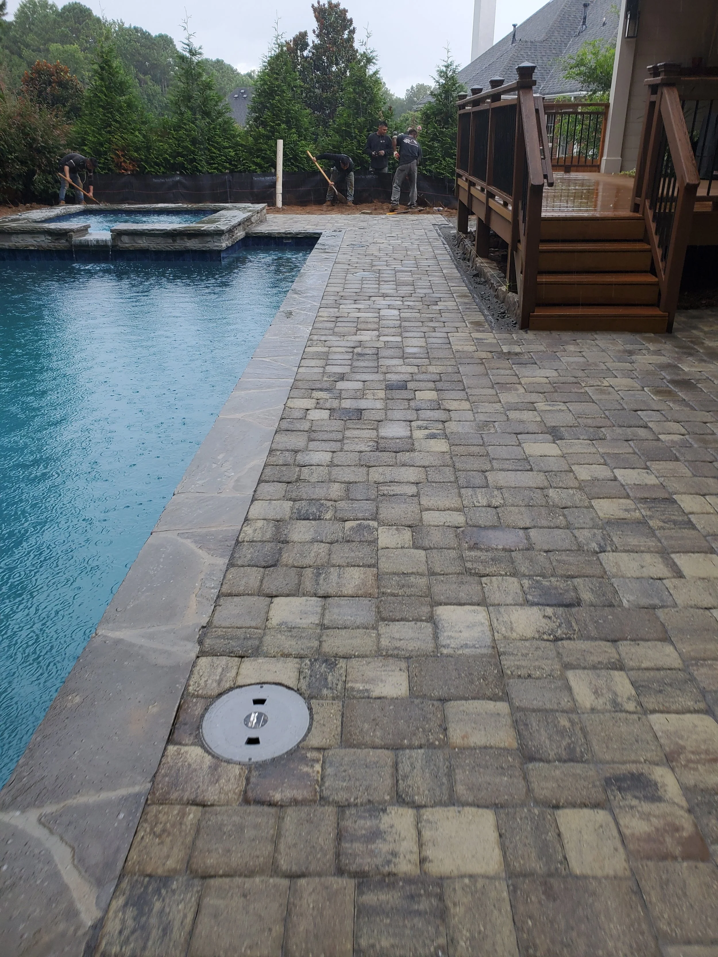 Workers installing or repairing a stone patio beside a swimming pool, with trees and a house in the background.