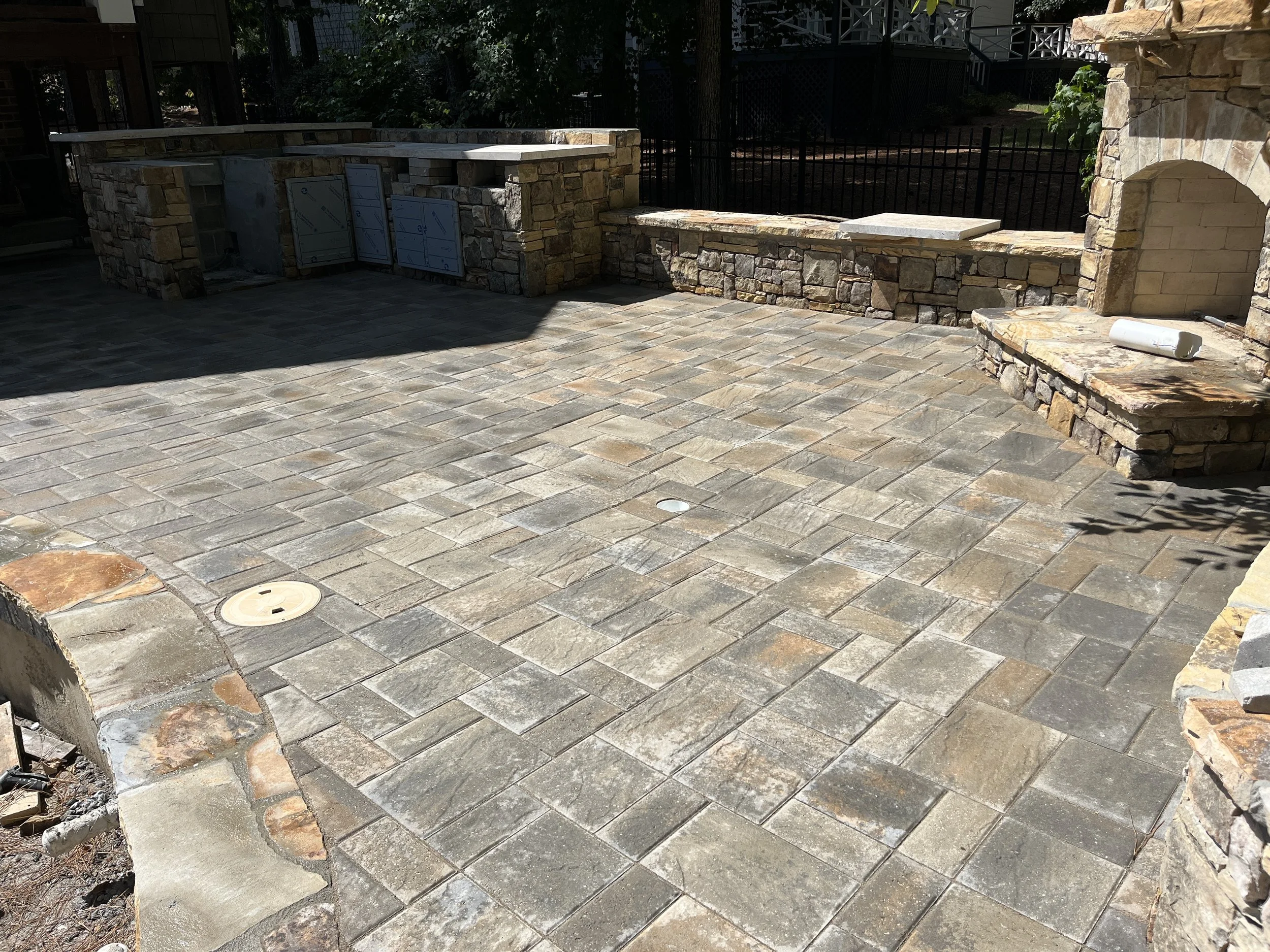 A patio area with stone pavers and a stone wall with an arch, a bench, and some construction materials. In the background, trees and a fencing are visible.