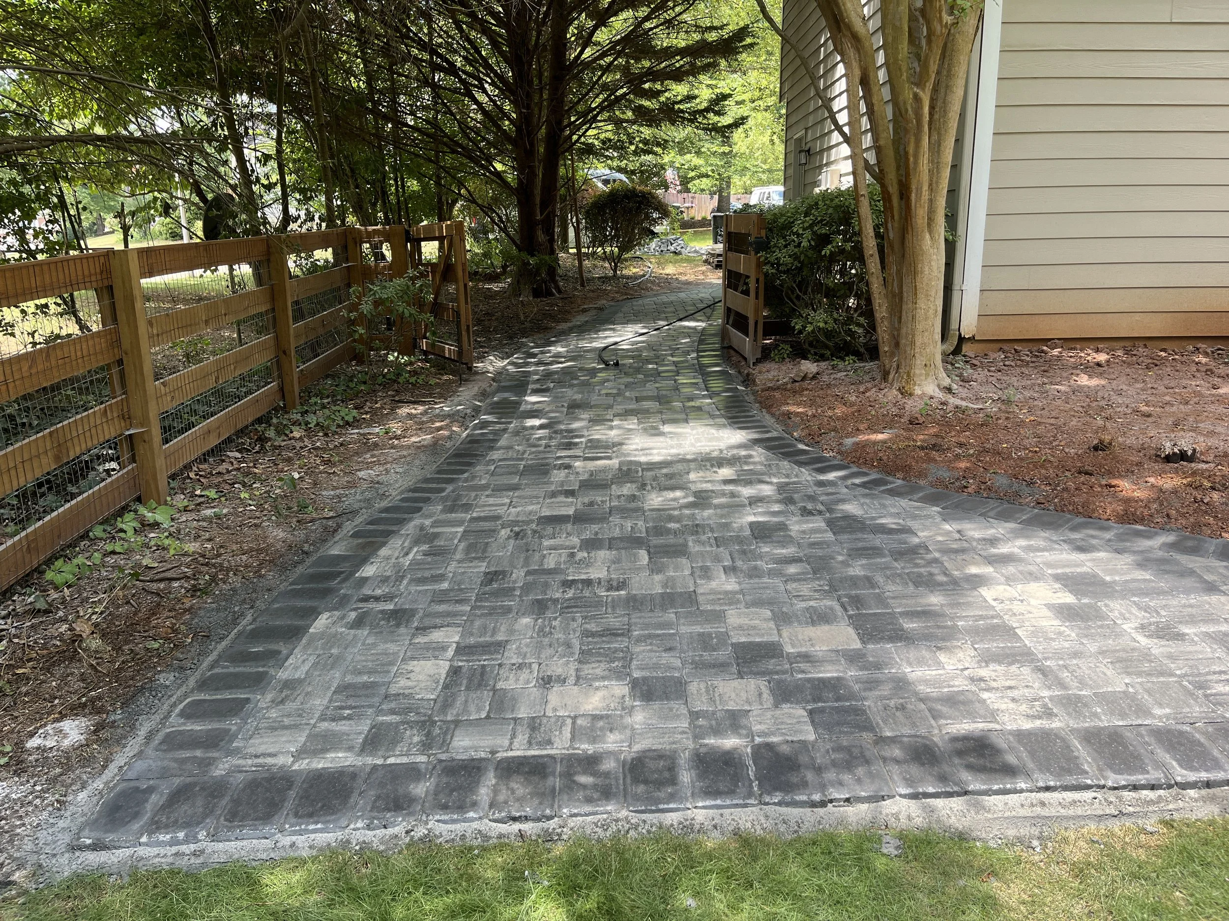 A newly paved winding brick walkway next to a beige house with exterior siding. There are trees and shrubs along the pathway, with a wooden fence on the left side and a garden area in the background.