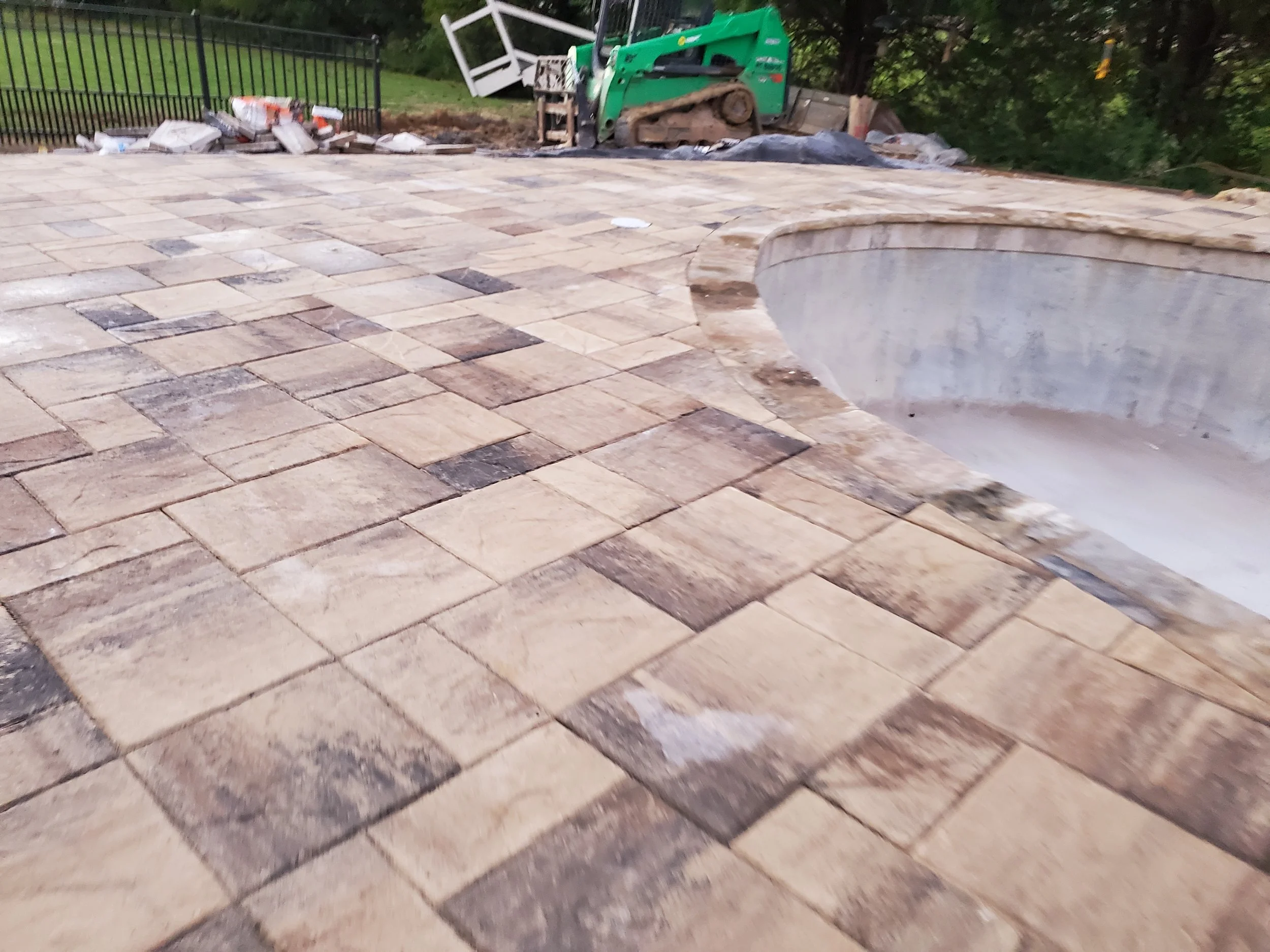 Newly paved stone patio with an empty swimming pool in the foreground, construction equipment, debris, and tools in the background