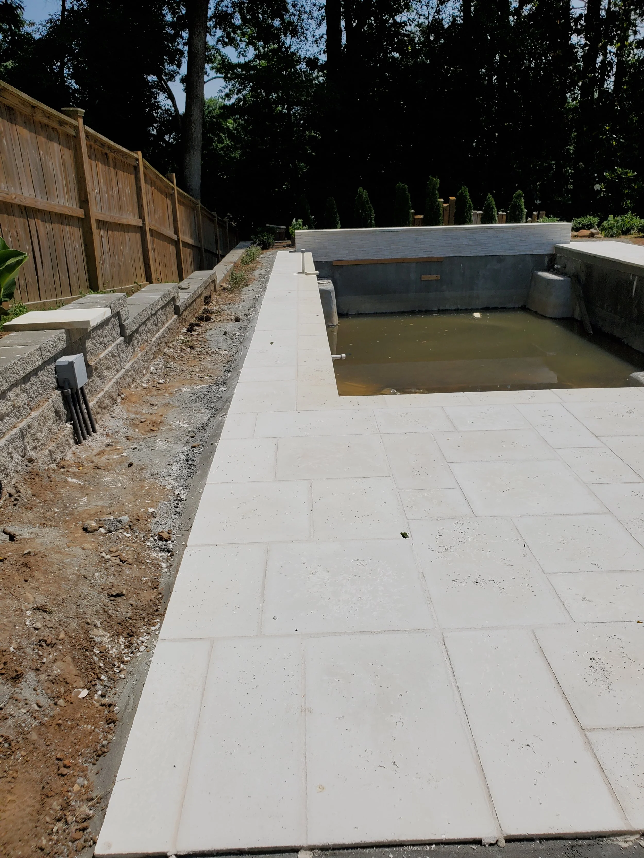 Under construction backyard with a small empty pool, white tile flooring, wooden fence, and tall trees in the background.