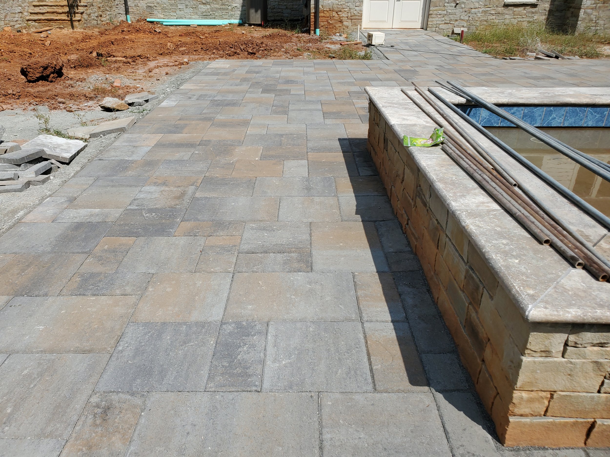 An outdoor area under construction with a partially completed paved patio, construction materials, and tools. The background features a brick wall, a door, and some exposed soil and grass.