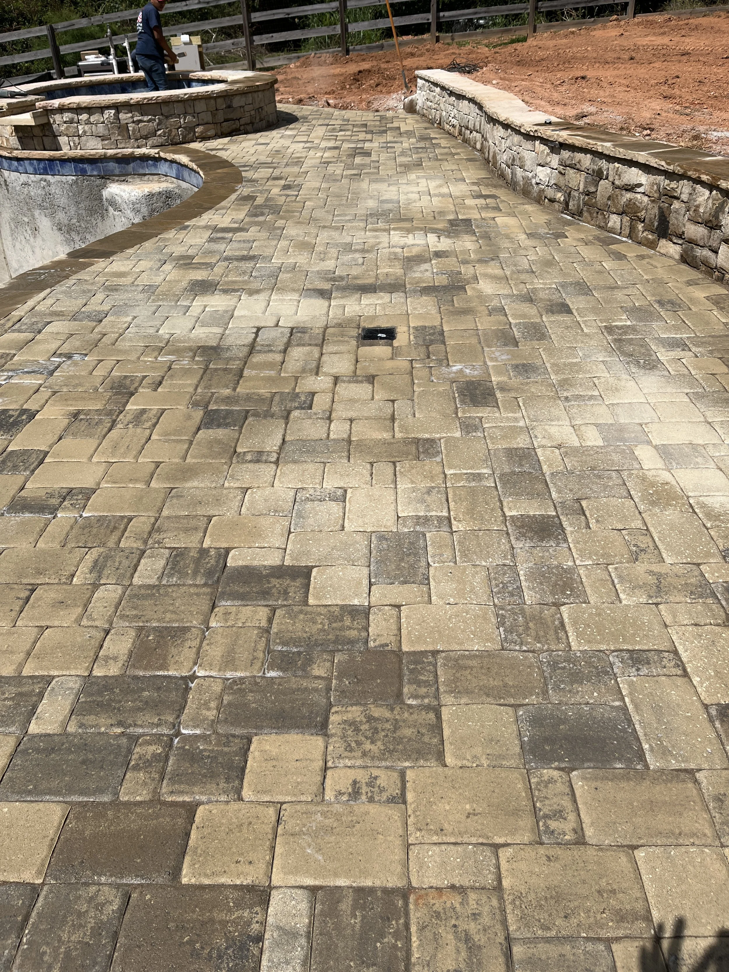 A newly paved stone patio with a curved edge, drainage grate in the center, stone retaining walls, and a hot tub under construction in the background, with a person working nearby.