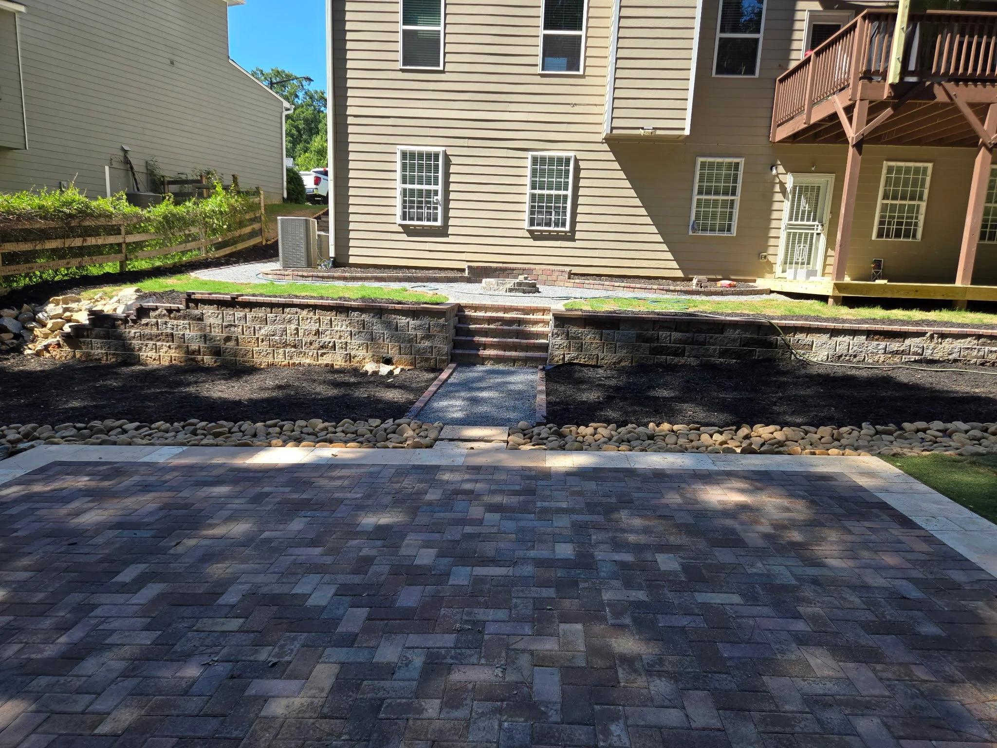 Backyard patio with brick pavers, retaining wall, small steps, grassy area, fenced garden, and part of house with beige siding and multiple windows.