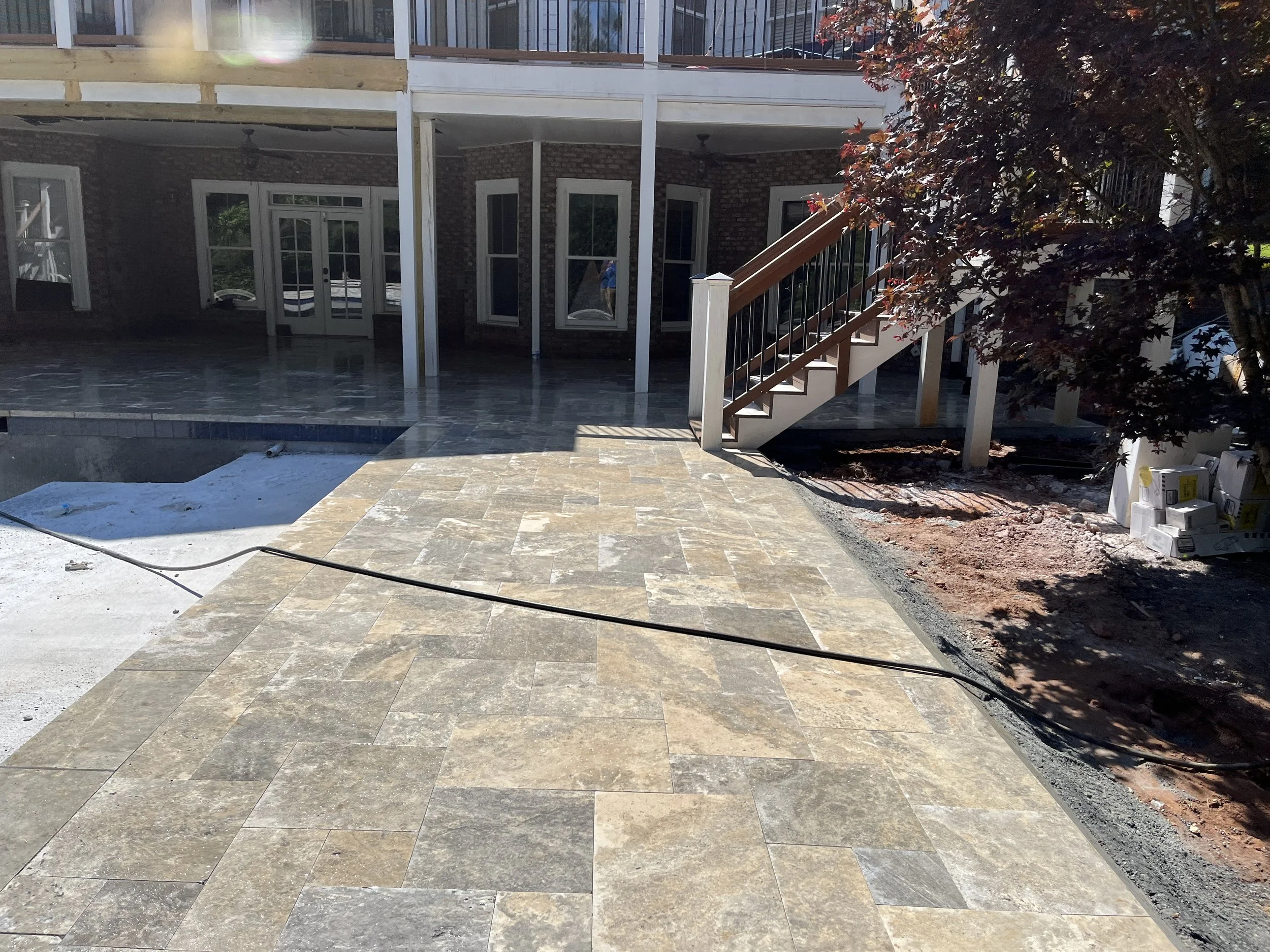 Backyard with a brick house, glass doors, a covered patio with a ceiling fan, and a staircase leading to an upper deck. The patio has stone flooring, and there are construction materials and tools visible on the side. A tree with red leaves partially