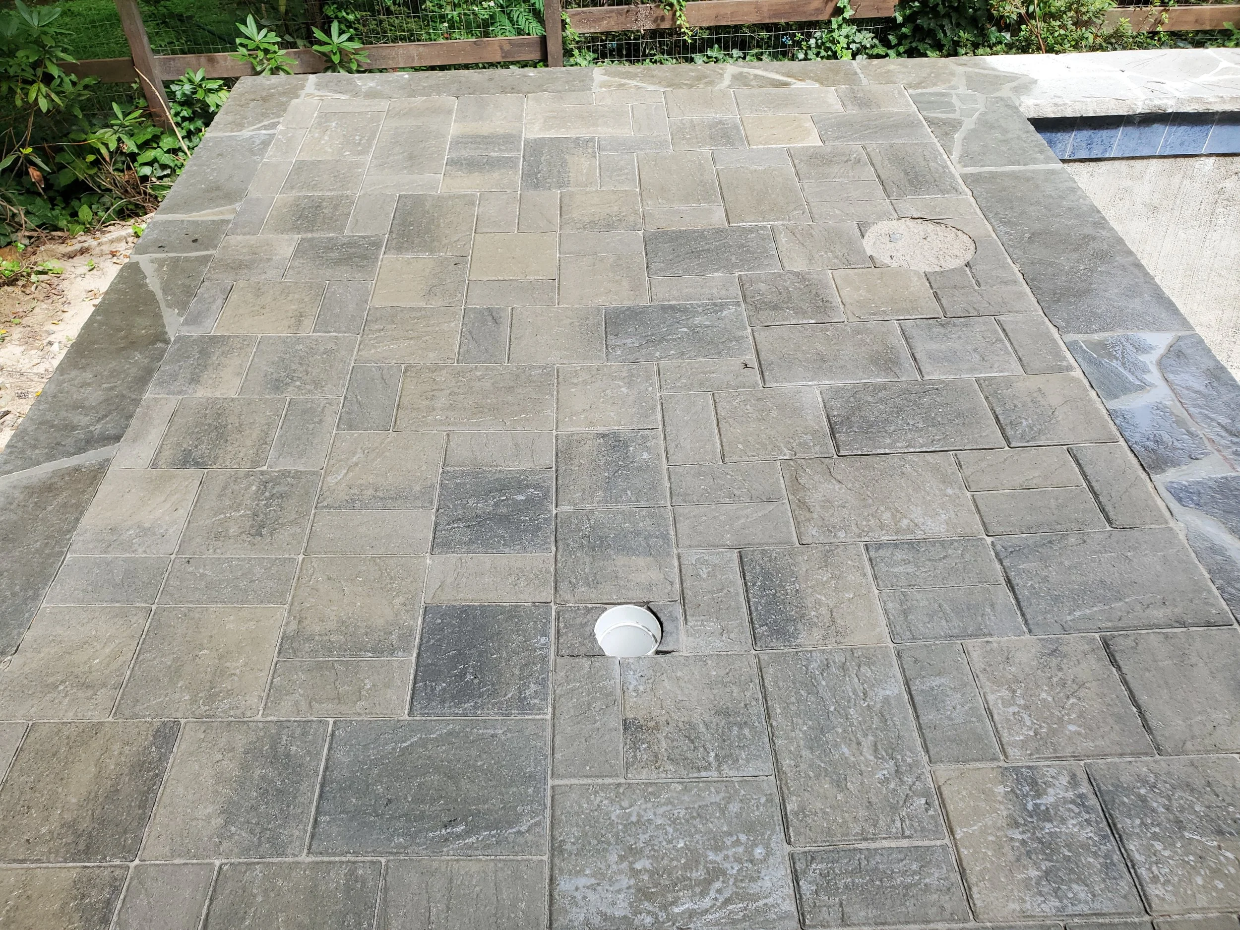 Outdoor area with gray stone tiled flooring, a wooden fence, green shrubbery, and a round open drain in the center of the tile.