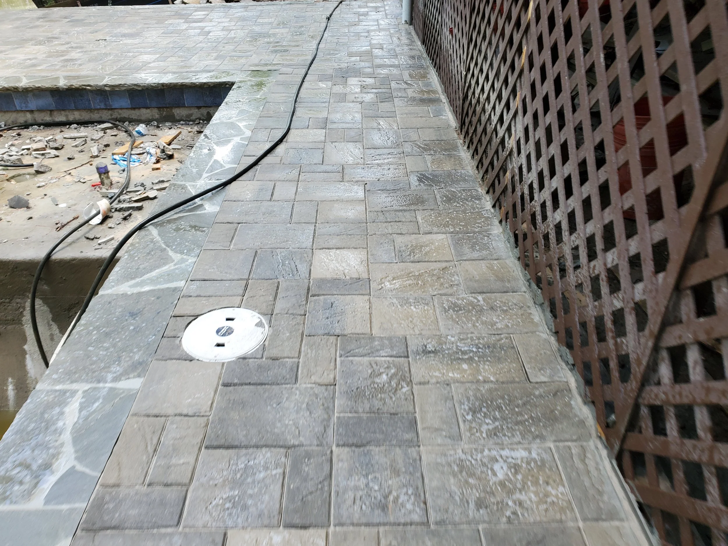 A partially constructed outdoor walkway with a new brick paver pattern, a drainage drain cover, construction debris, a black cable, and a wooden lattice fence on the right side.