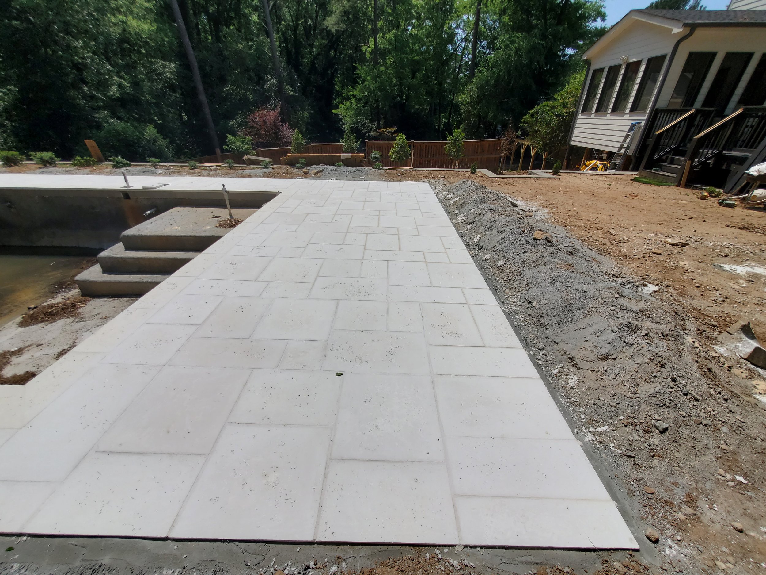 Construction site with newly laid white stone pathway, wooden fence, and a white house with a deck, surrounded by trees.