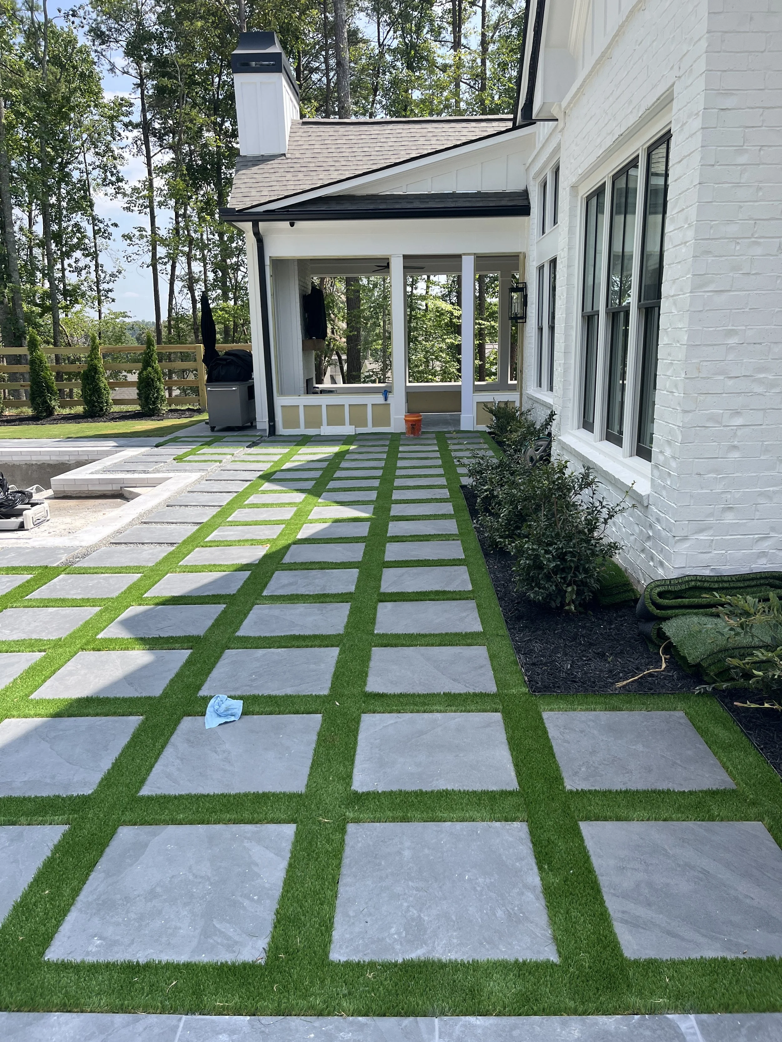 The image shows a backyard patio with a checkered pattern of large square concrete pavers and green grass strips. There is a white brick house with multiple windows on the right. The patio area has a small shrub and some other plants along the house,