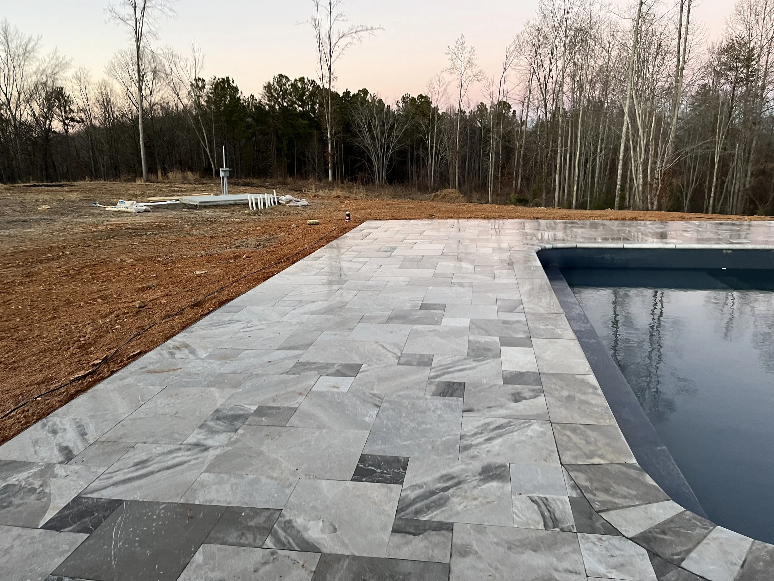 A partially constructed backyard with a newly installed stone patio or pool deck, surrounded by bare soil and trees in the background, during dusk or dawn.