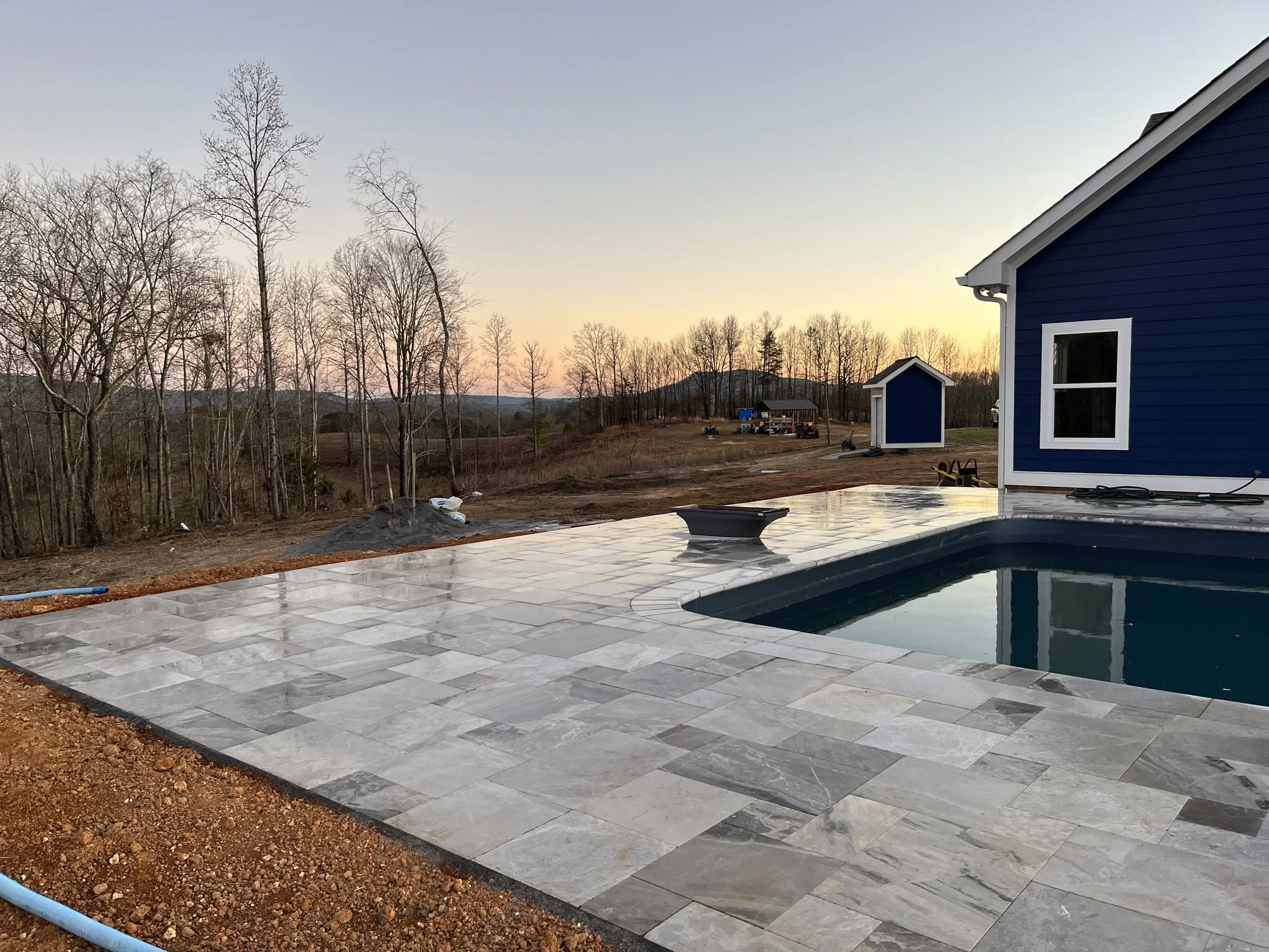 A backyard scene during sunset showing a partially completed pool area with a tiled deck, a blue house on the right, and leafless trees in the background.