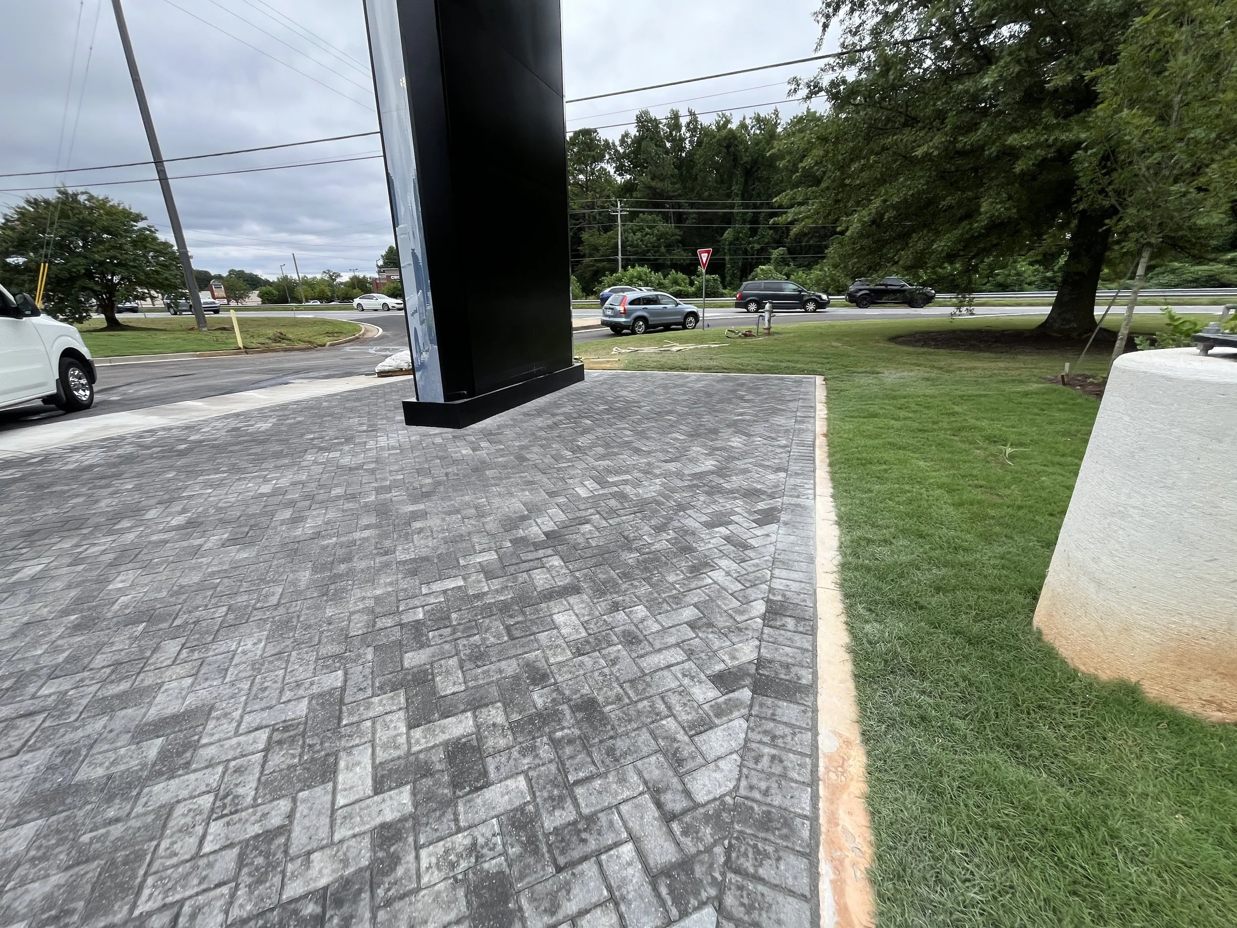A parking lot area with a brick-paved sidewalk, several cars, a fire hydrant, traffic, and trees under a cloudy sky.