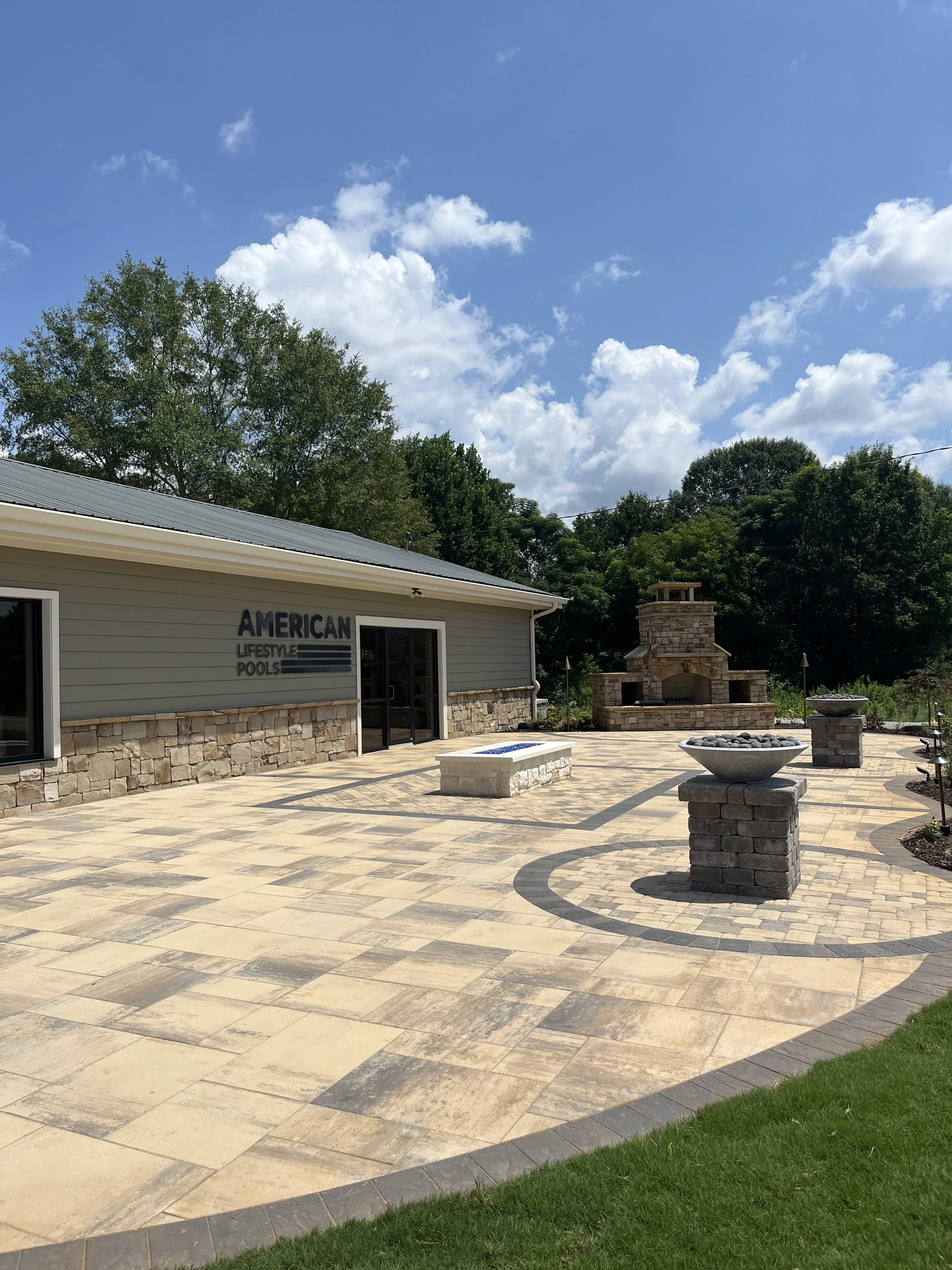 A patio with patterned stone tiles, outdoor fireplace, and a building with a sign that reads 'American Lifestyle Pools.' There is a grassy area in the foreground, and trees and a blue sky with clouds in the background.