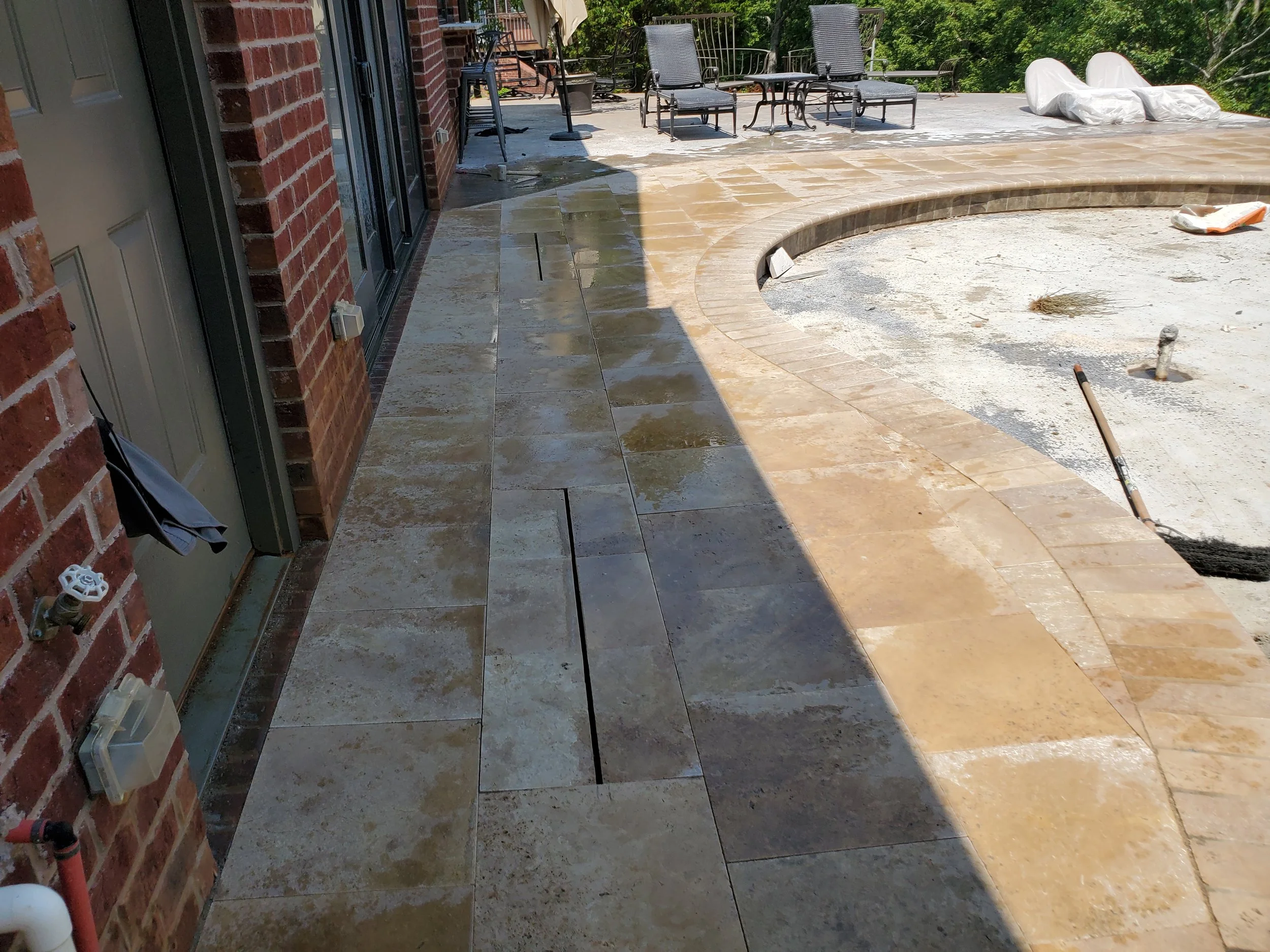 Backyard patio under construction with partially installed large beige tiles, outdoor furniture, and construction tools and materials.