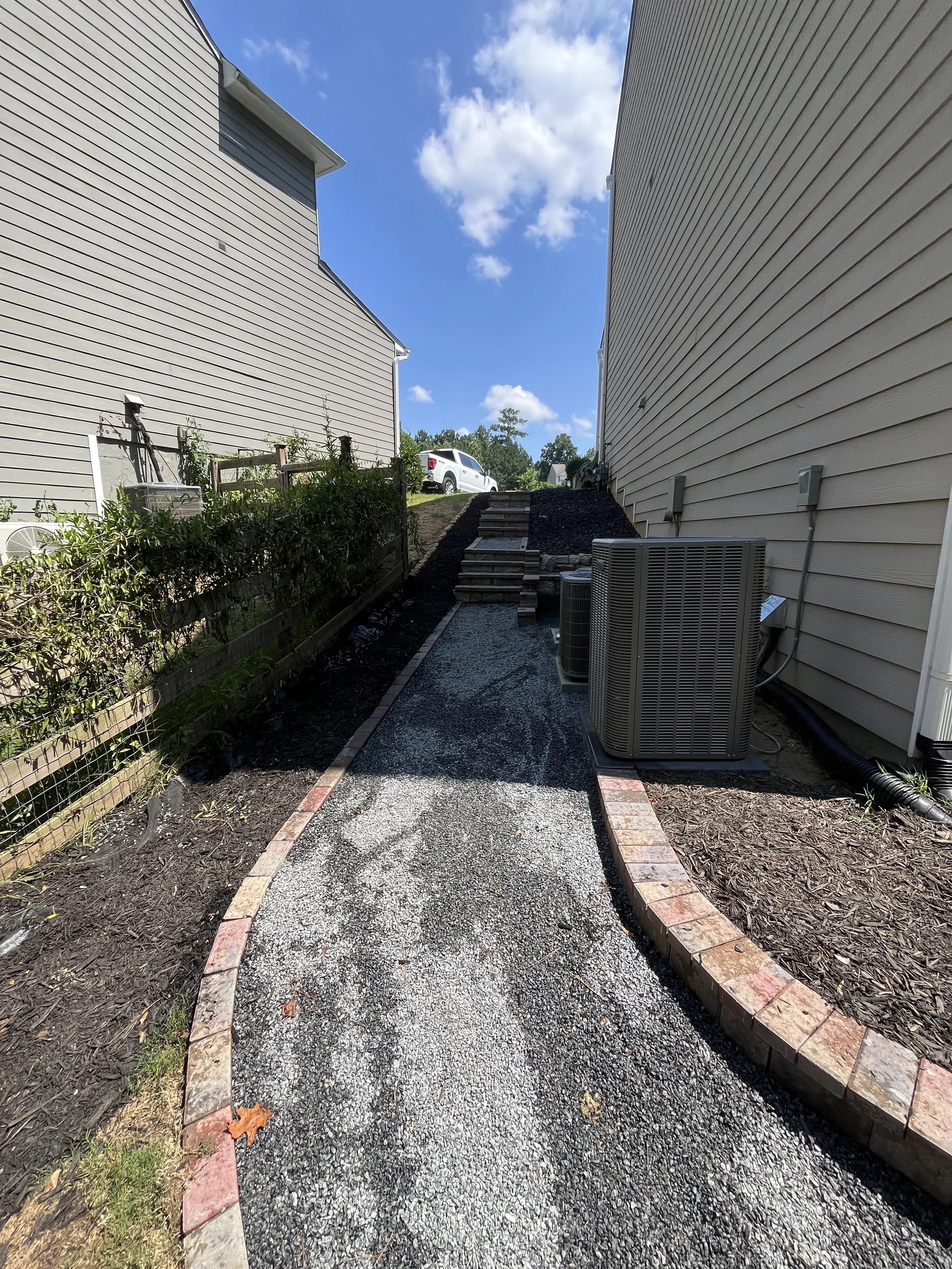 A narrow gravel pathway between two beige houses with siding, bordered by bricks, leading to stairs and a white pickup truck in the distance under a partly cloudy sky.