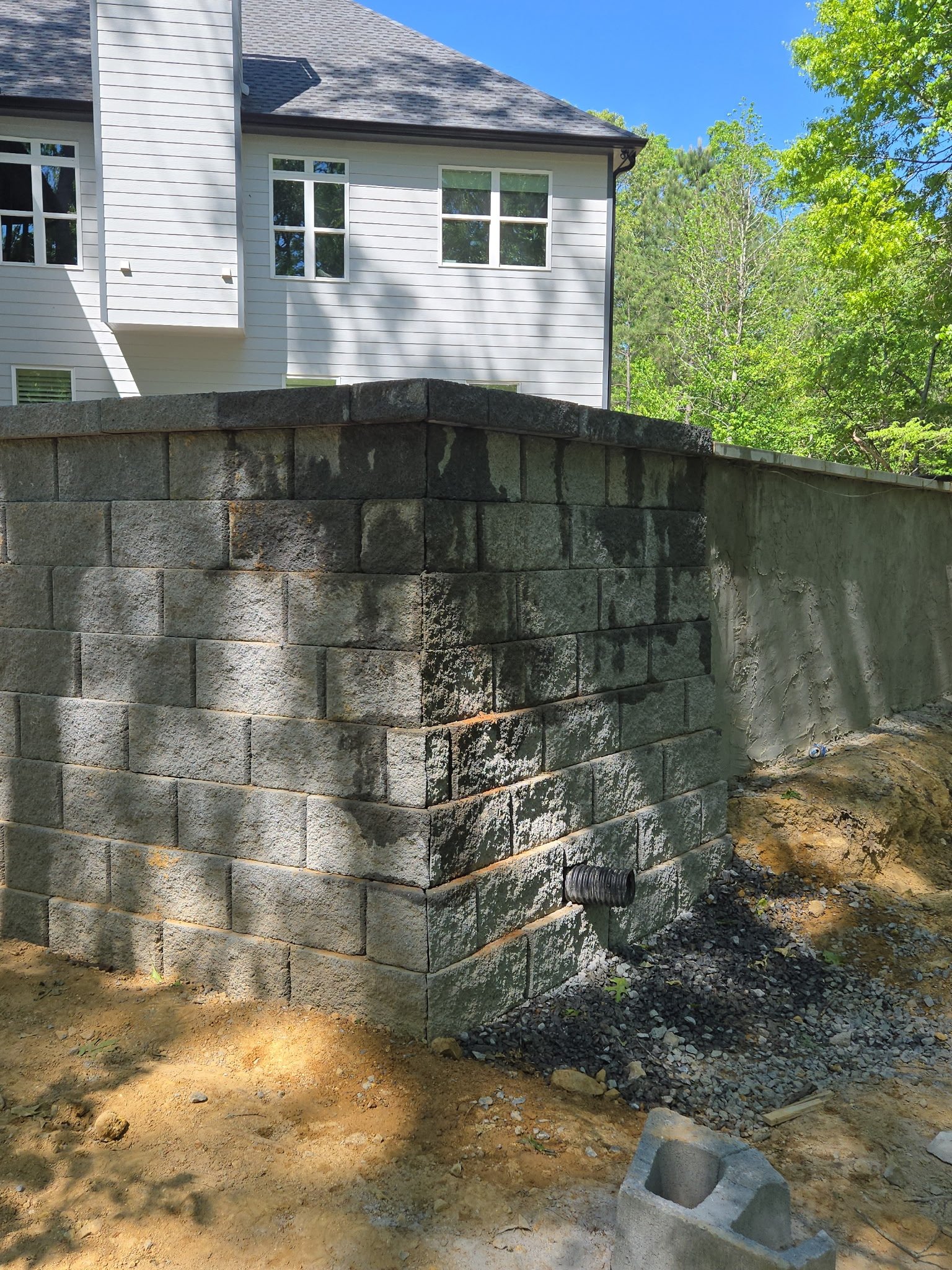 Construction site with a partially built cinder block wall in front of a white house with large windows, surrounded by trees, with a clear blue sky overhead.
