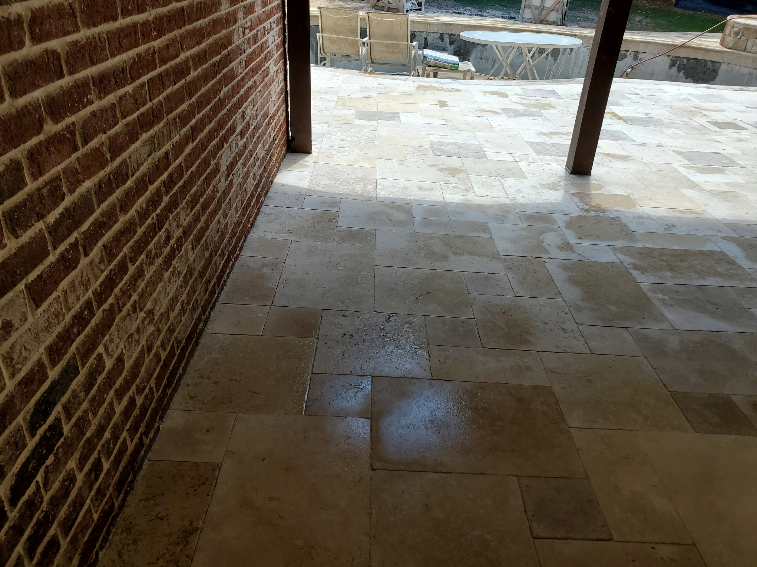 View of a patio with beige stone tile flooring, partial brick wall on the left, and outdoor furniture including chairs and a table in the background. There is a pool or water feature and surrounding area visible beyond the patio.