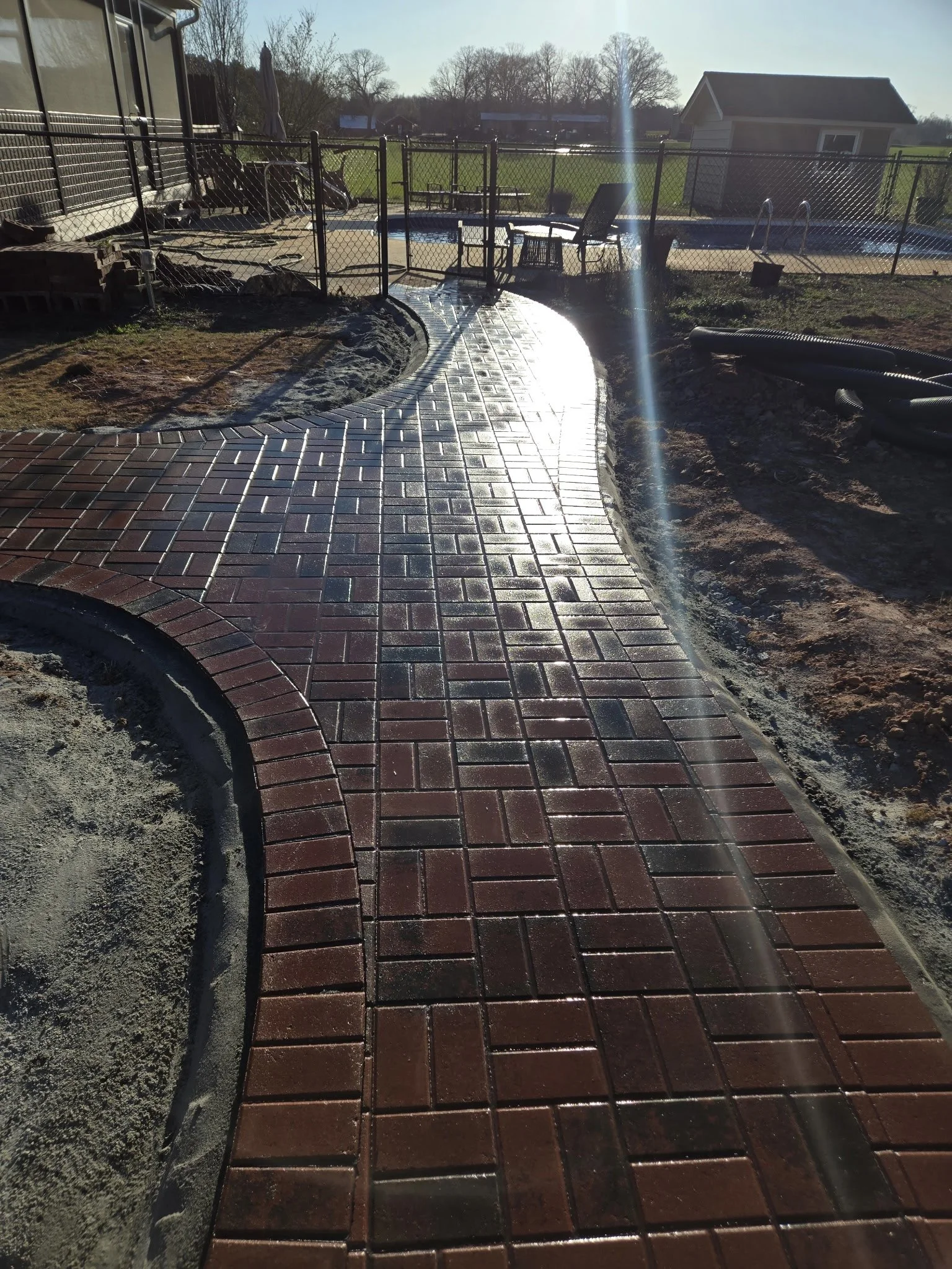 A backyard with a new brick pathway leading to a fenced pool area. The pathway curves and has dark red bricks with a glossy finish. The pool is behind a black chain link fence, with outdoor furniture and a small shed visible in the background. The ya