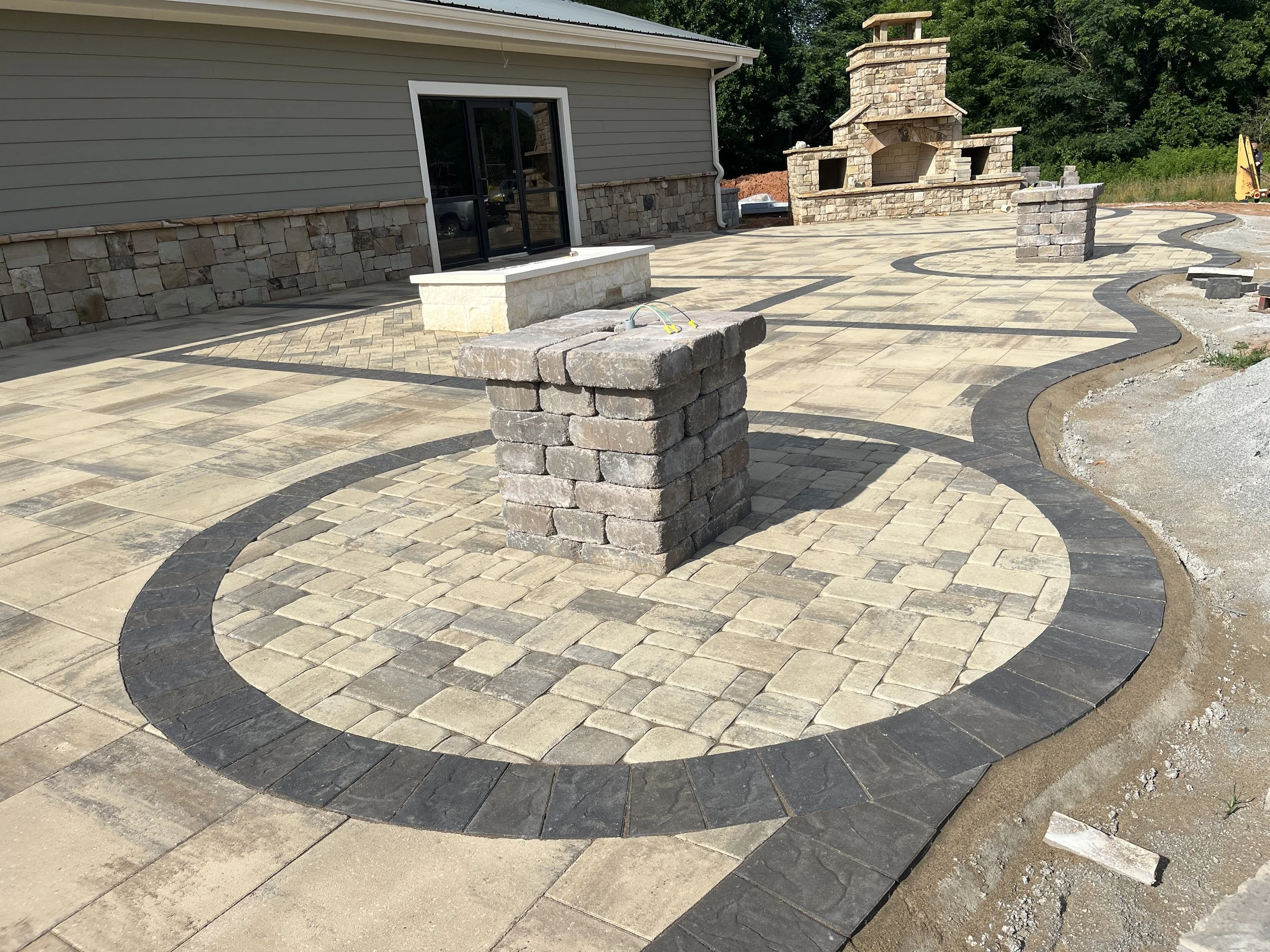 A backyard patio with decorative stone pavers, a curved border of darker stones, a brick and stone outdoor fireplace, and a green landscape with trees in the background.
