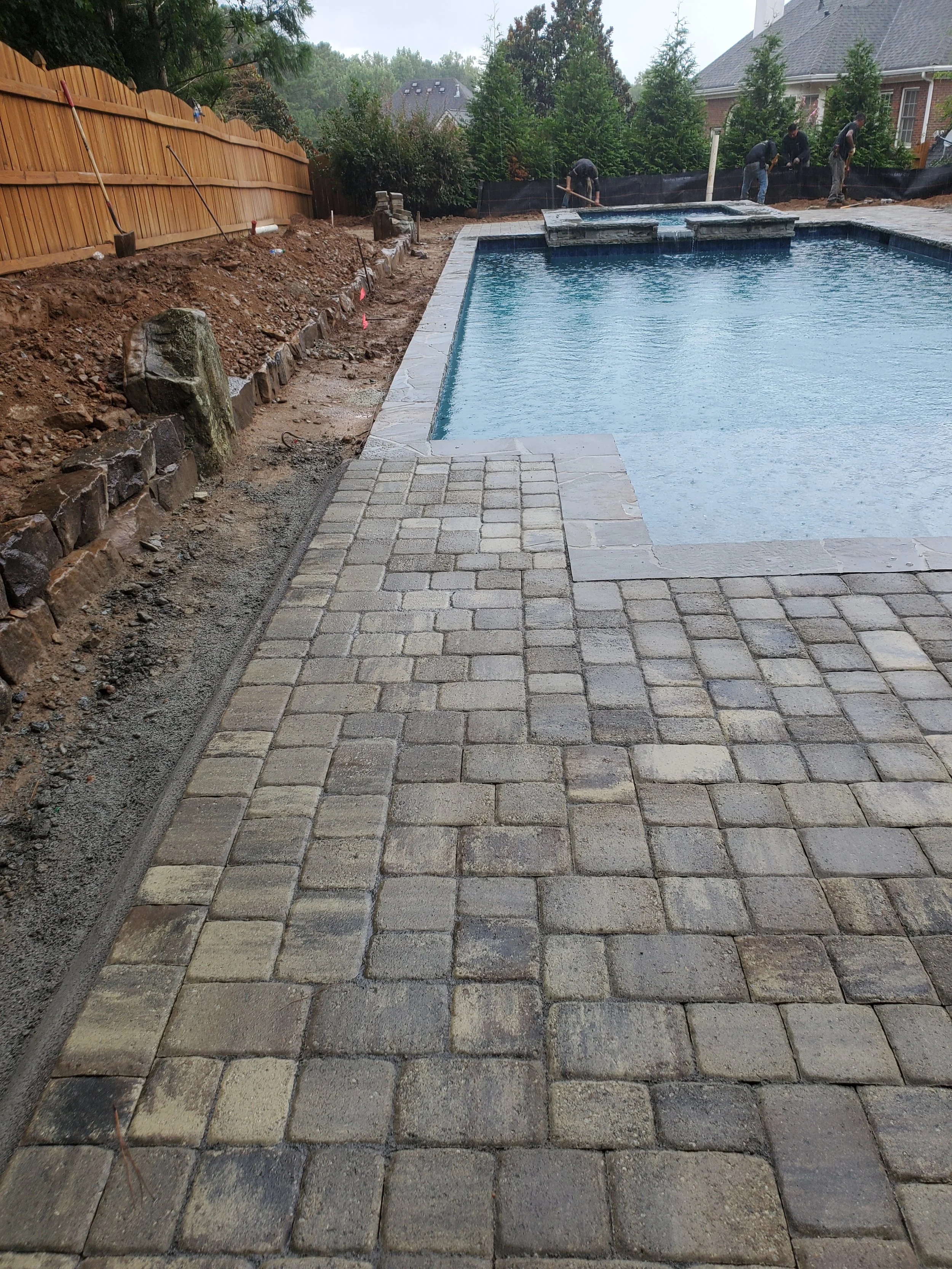 Backyard pool under construction with workers, wooden privacy fence on the left, and a stone patio in progress.
