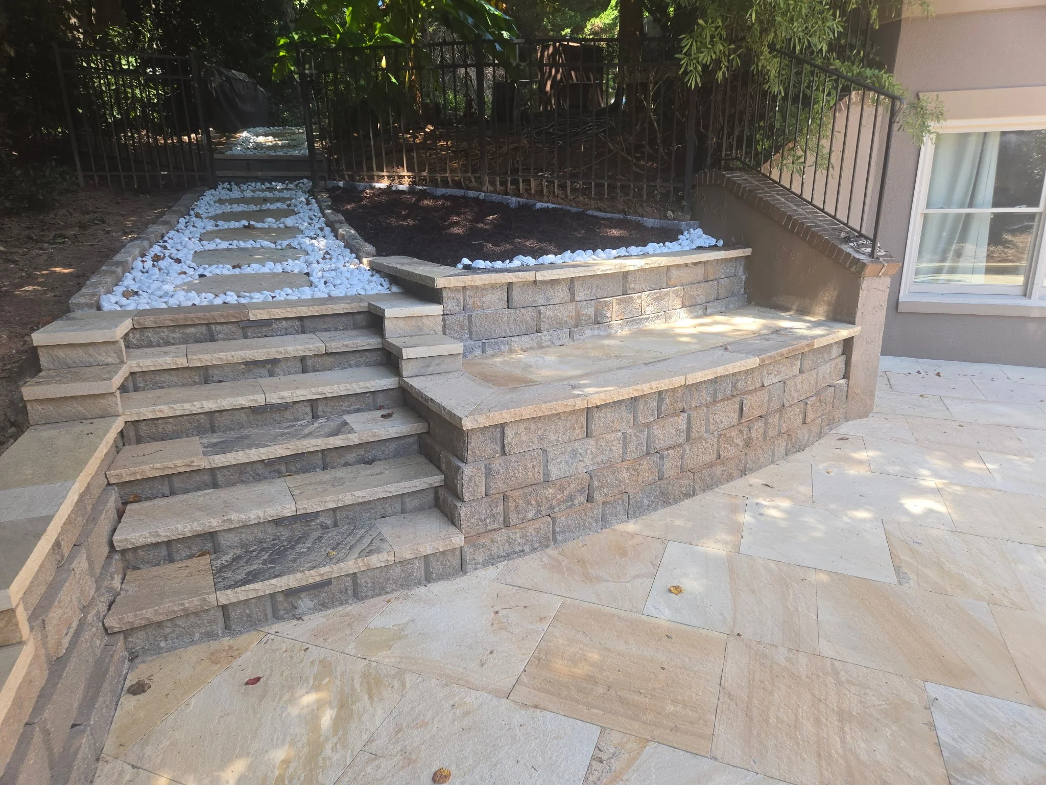 Stone steps leading up to a small garden bed surrounded by white rocks, with a black metal fence and lush green trees and plants in the background.