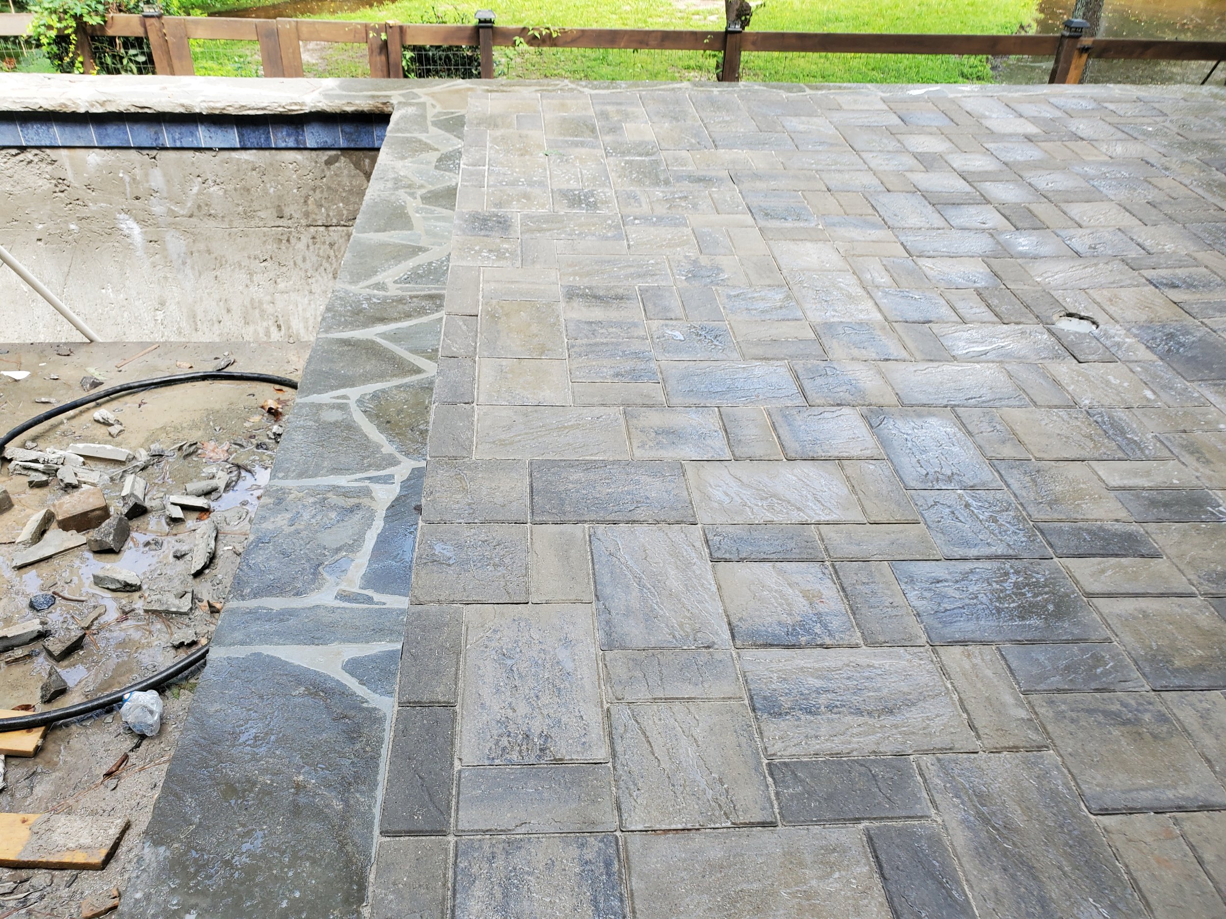Partially completed outdoor stone patio with bricks and stonework, adjacent to a raised bed with a wooden and wire fence, and a grassy backyard in the background.