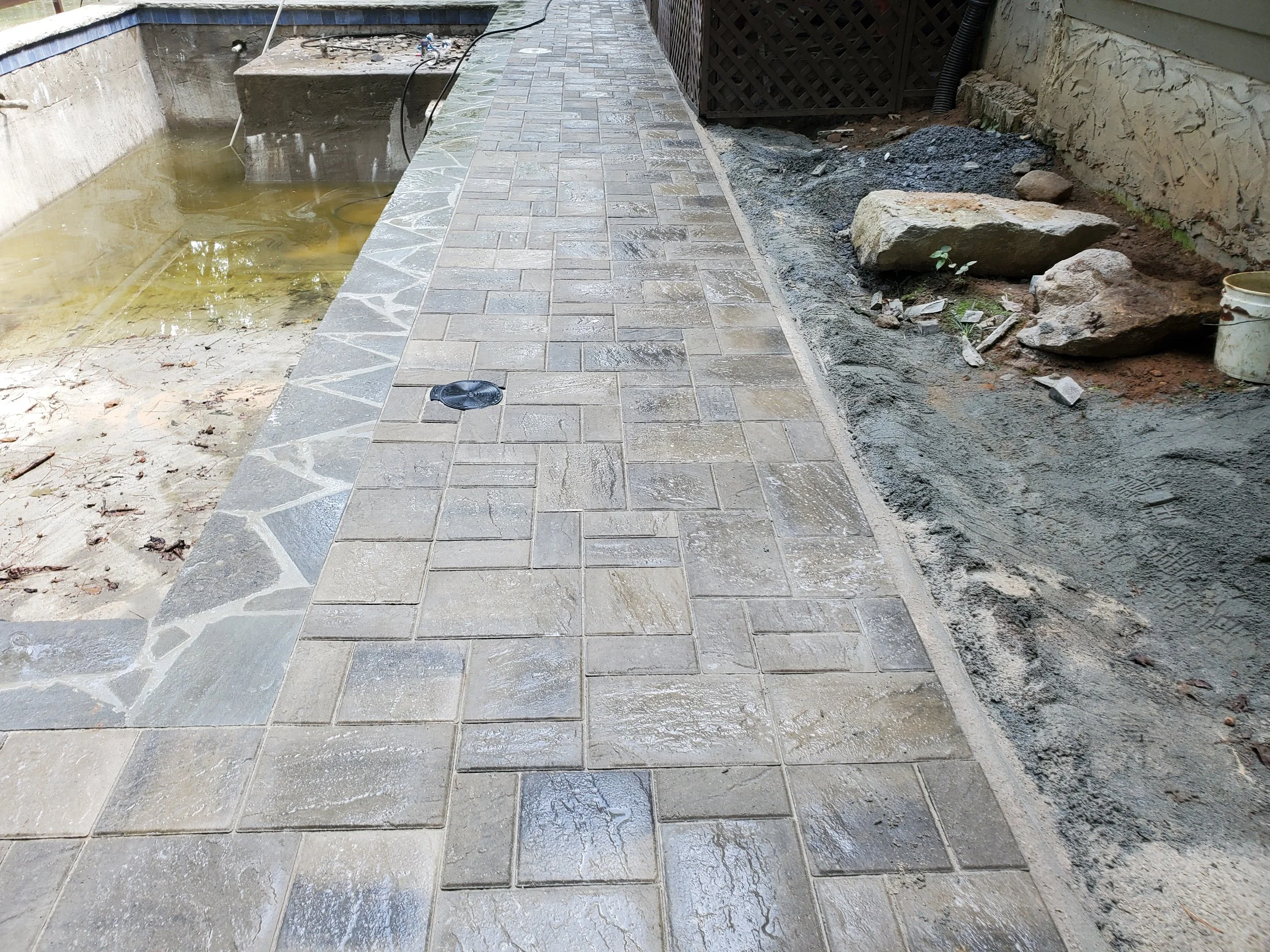 A partially finished outdoor patio with newly installed beige and gray patterned stone tiles. To the right, there is an area with loose dirt, rocks, and some small plants, indicating ongoing garden or landscaping work. A large rock and a bucket are a