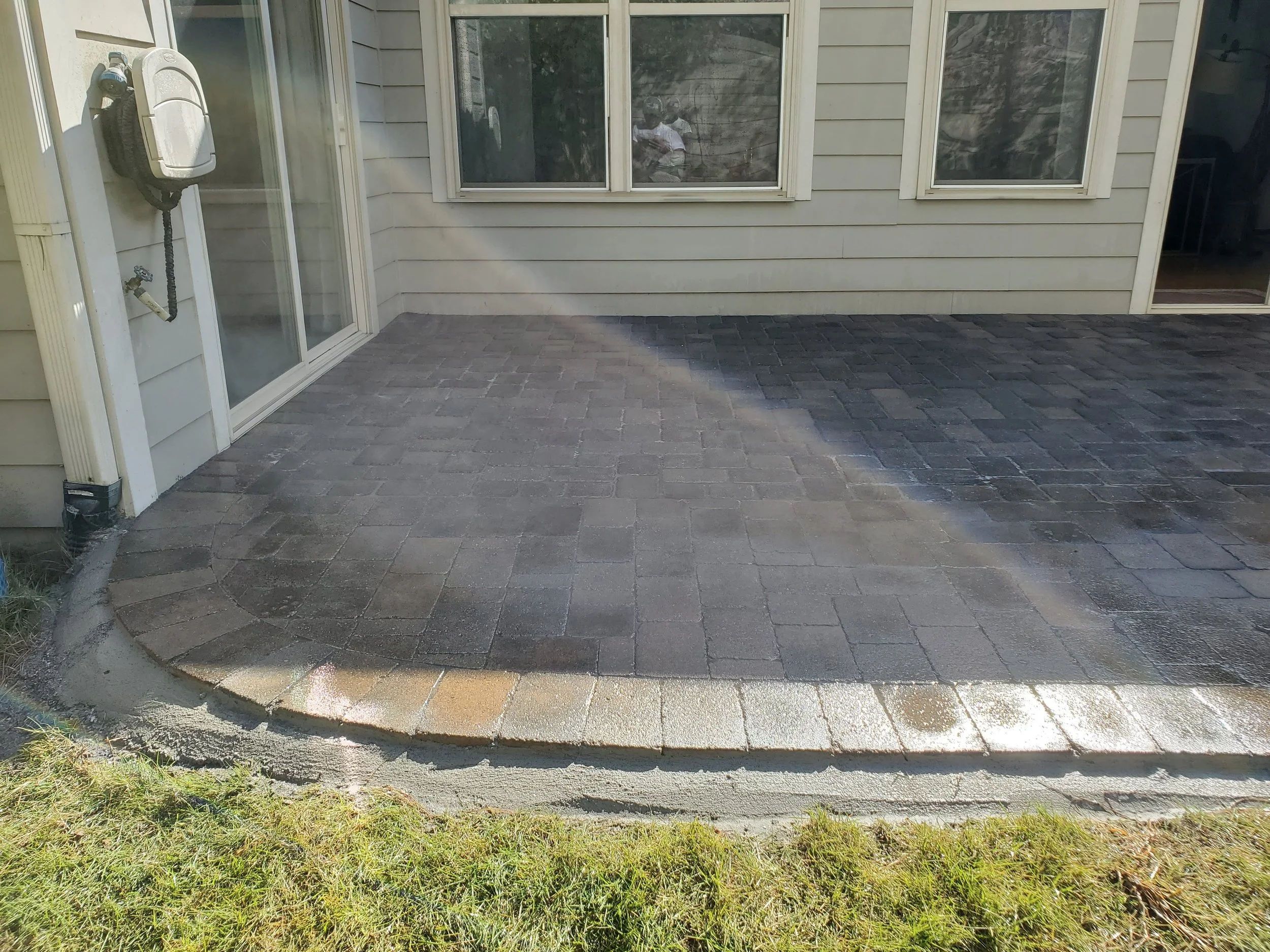 A newly paved brick patio with a curved edge, adjacent to a beige house with siding and large sliding glass doors. There is a grassy area in the foreground and a reflection of a person in the patio door window.