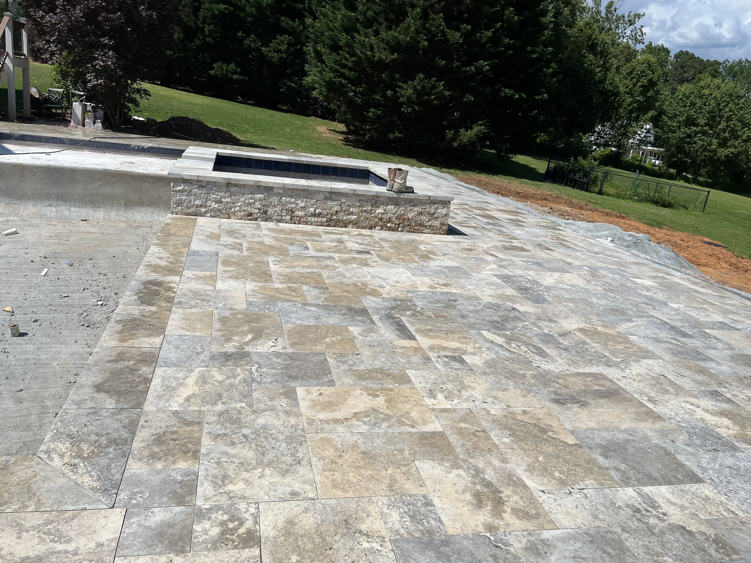 Construction site with a partially finished stone patio and a raised hot tub, surrounded by a grassy yard with trees in the background.
