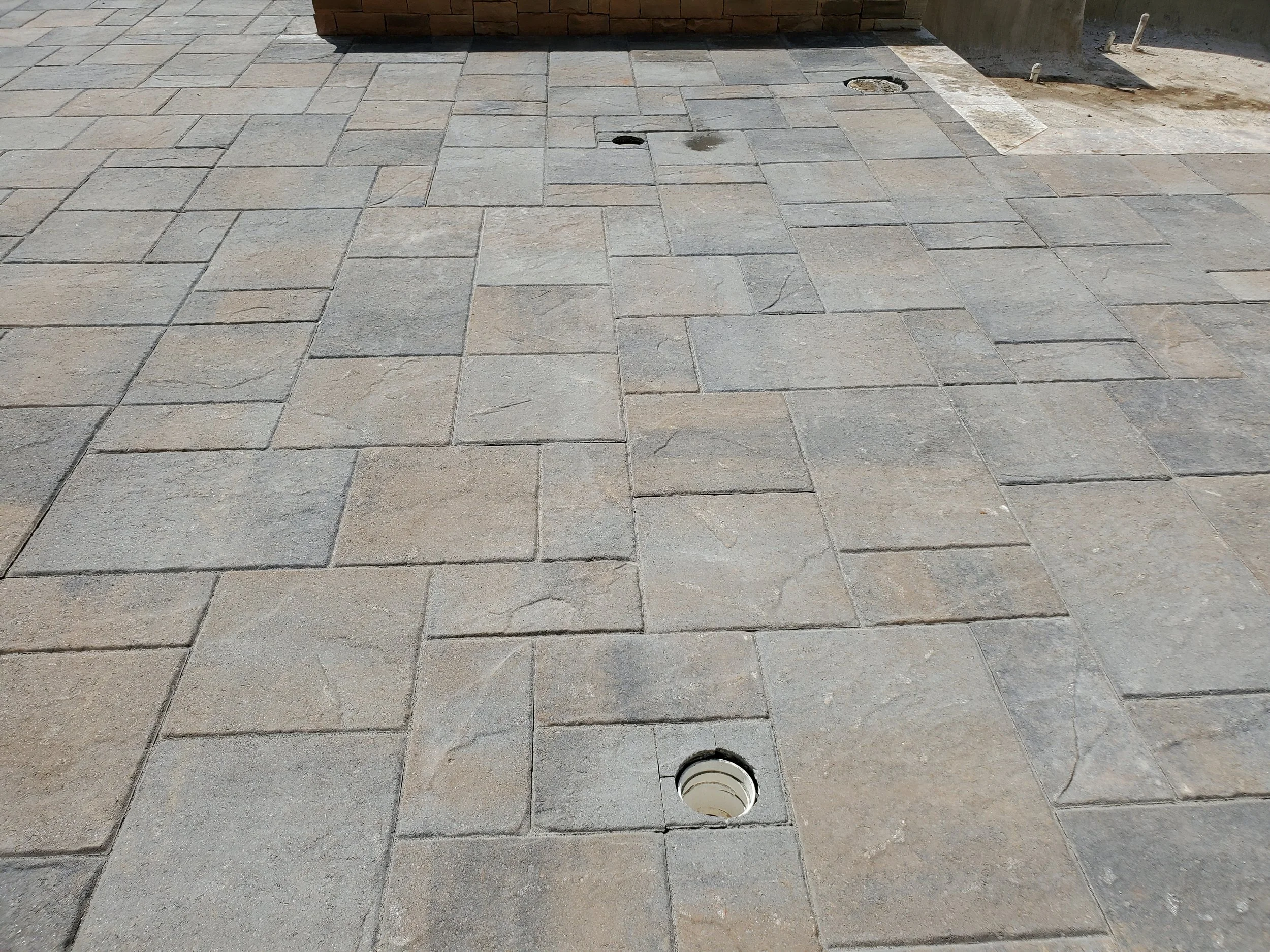 Paved sidewalk with two circular holes, one near the top center with a black cover, and the other near the bottom center with a white cover, next to a brick wall and a small patch of exposed dirt and concrete