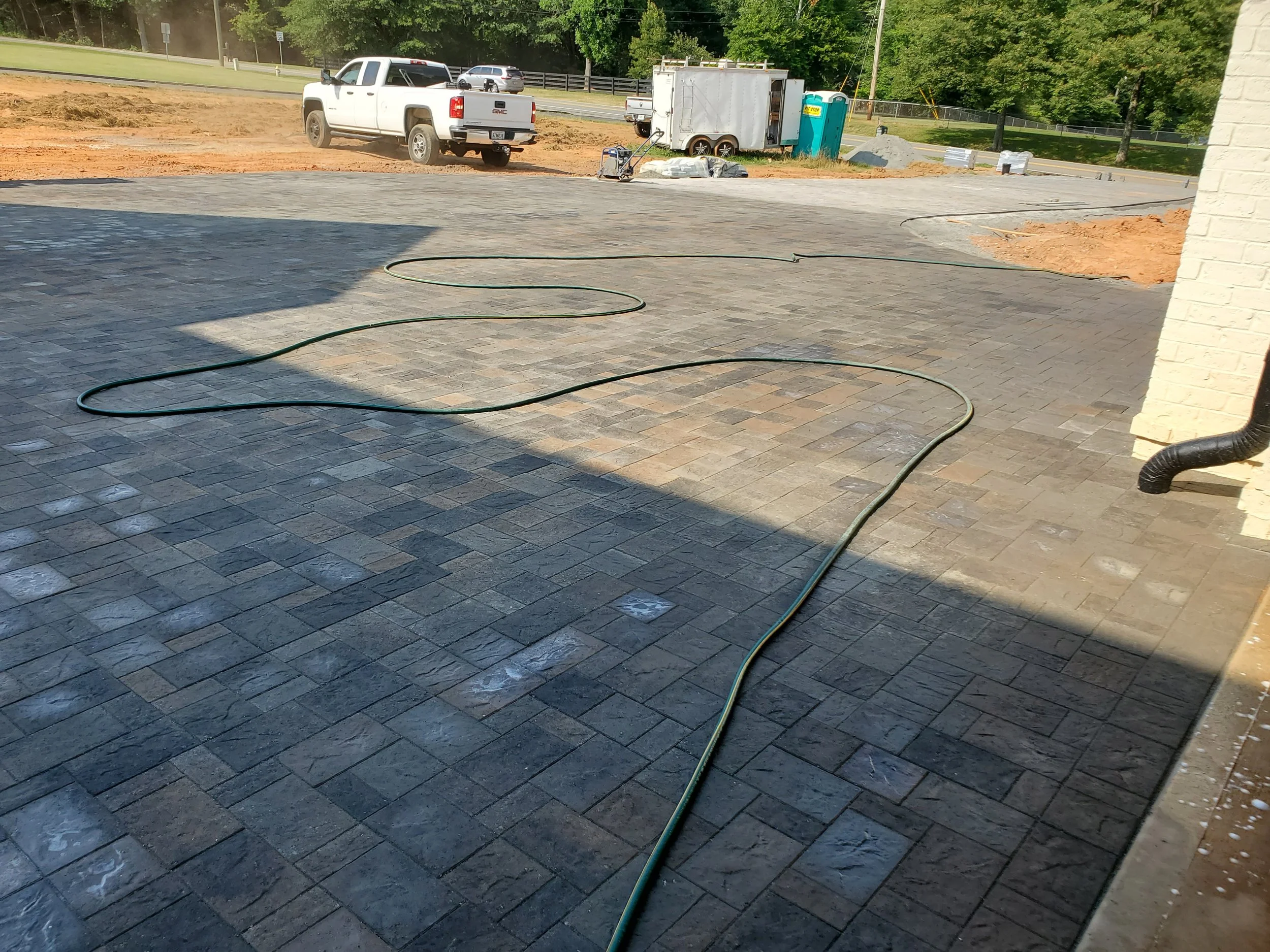 Construction or landscaping site with a partially paved area, a garden hose on the ground, a white pickup truck, a construction trailer, a portable toilet, and construction materials in the background.