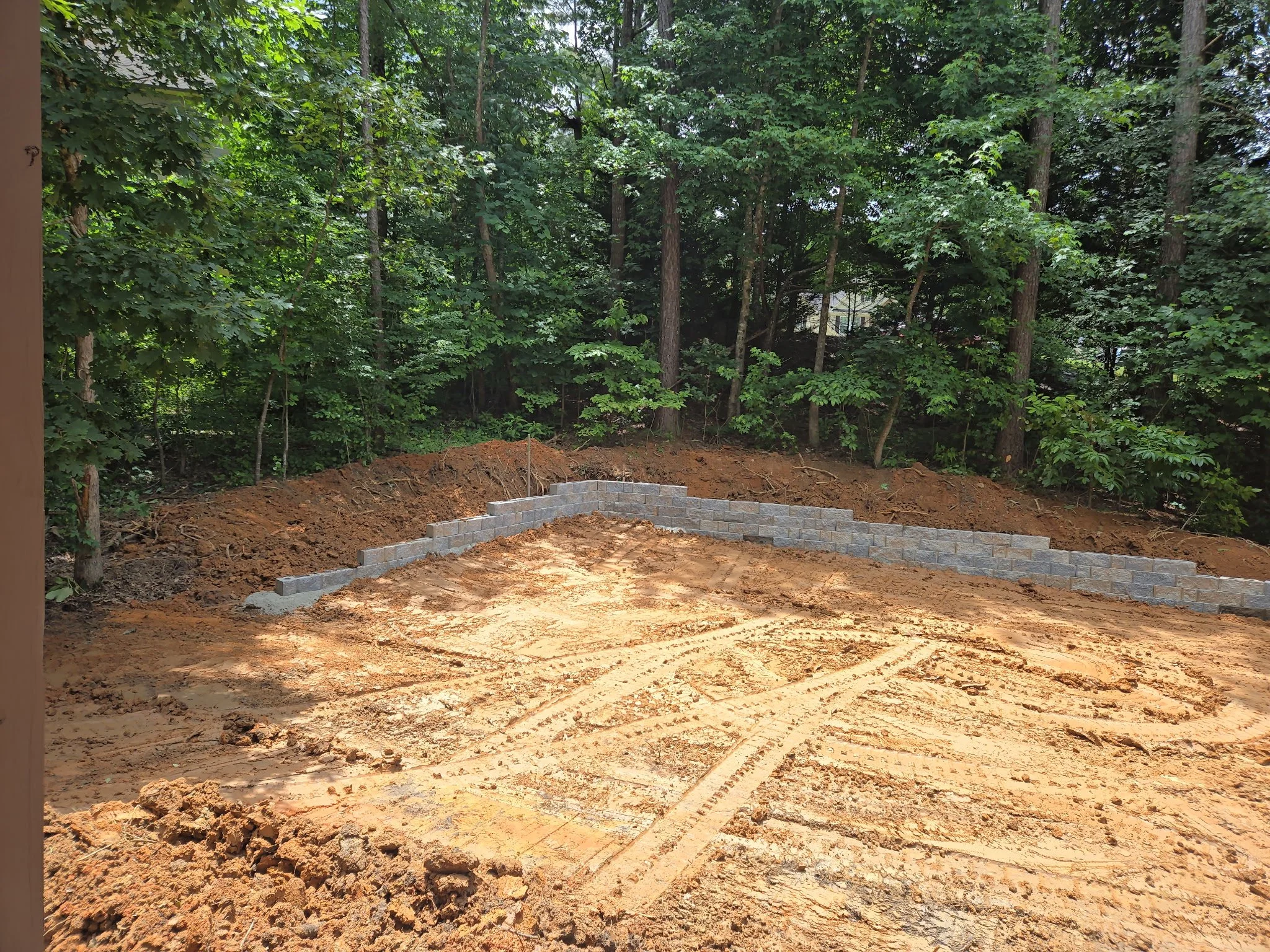 A construction site in a wooded area with a partially built stone wall and exposed dirt ground.