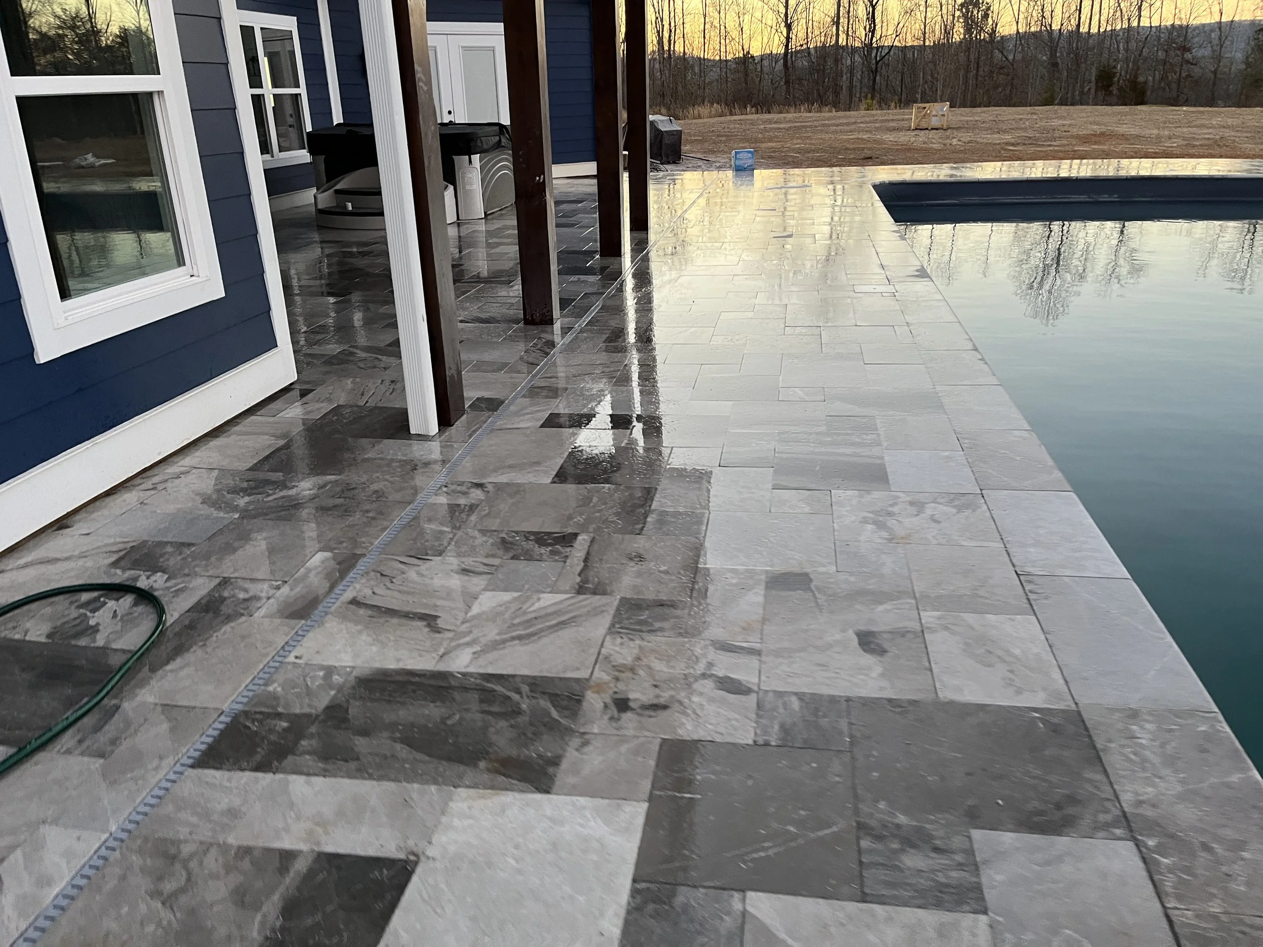 Backyard patio area with tiled flooring, a pool, and a house with blue siding and white trim; outdoor sunset scene with trees and mountains in the background.