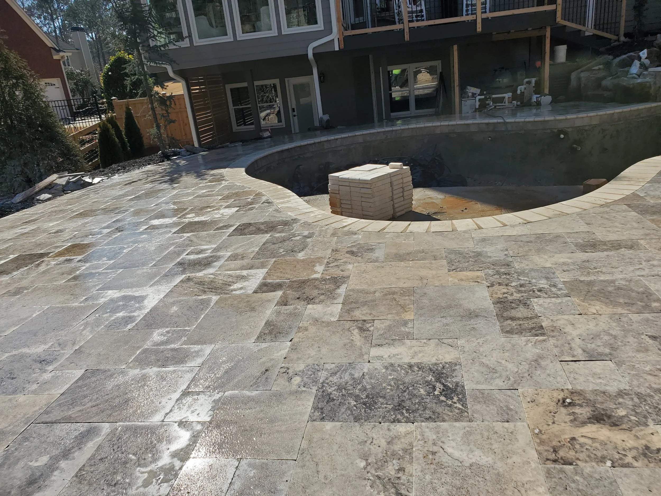 A backyard with a stone patio, a swimming pool under construction with layers of bricks on the edge, and a house with a deck in the background.
