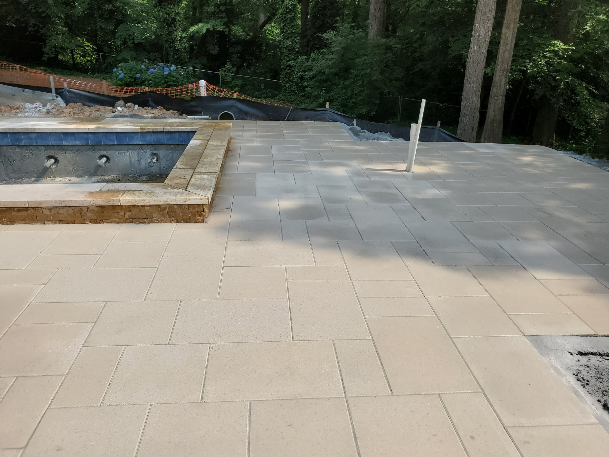 An outdoor area under construction with light-colored tiled flooring, a small raised pool or hot tub with water jets, and a landscaped background with trees and bushes. An orange safety fence and construction materials are visible.