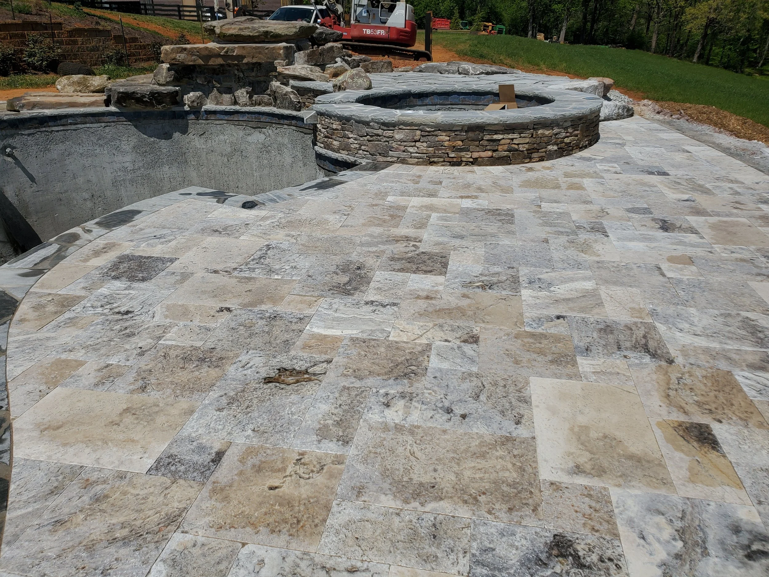 Construction site featuring a partially completed outdoor pool with stone and tile edging, a stone wall, a pile of large rocks, and construction equipment in the background.
