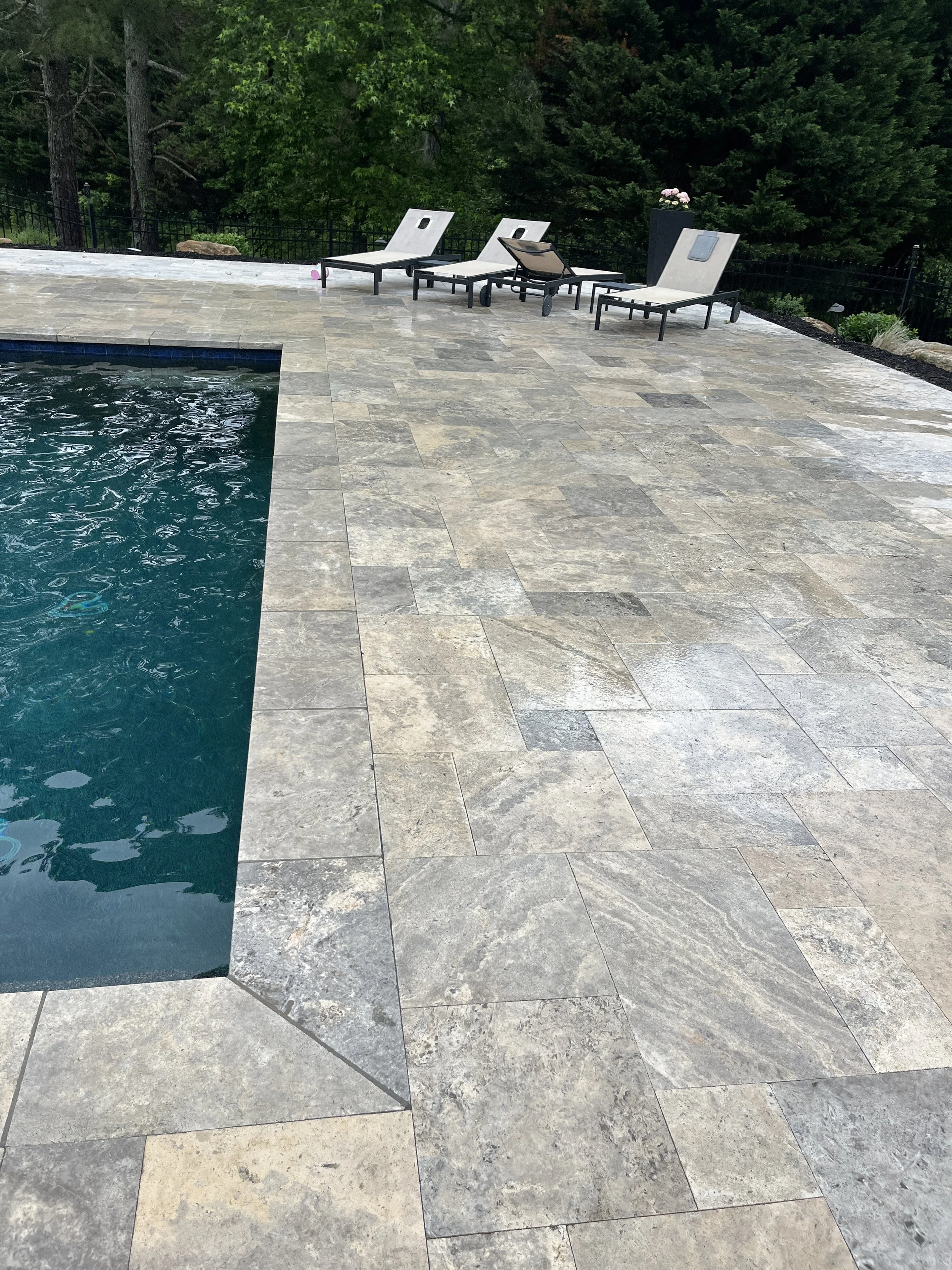 A backyard with a swimming pool and four lounge chairs on a stone deck, with a black fence and green trees in the background. One lounge chair is tilted back.