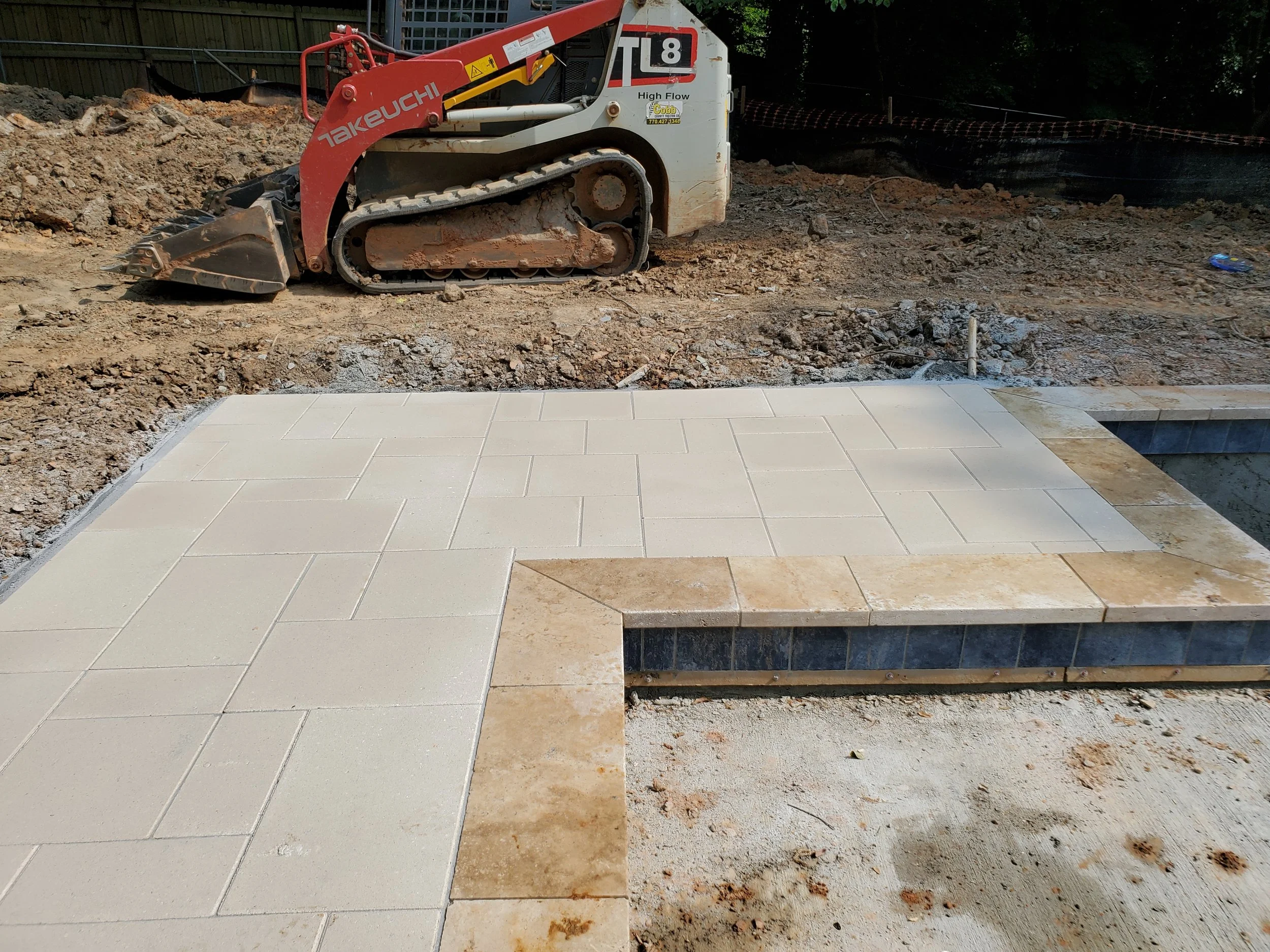 A construction site with a small dirt track loader and a partially finished tiled pool. The tiled pool has beige and dark blue tiles, and the background shows excavated soil and trees with a black plastic barrier.