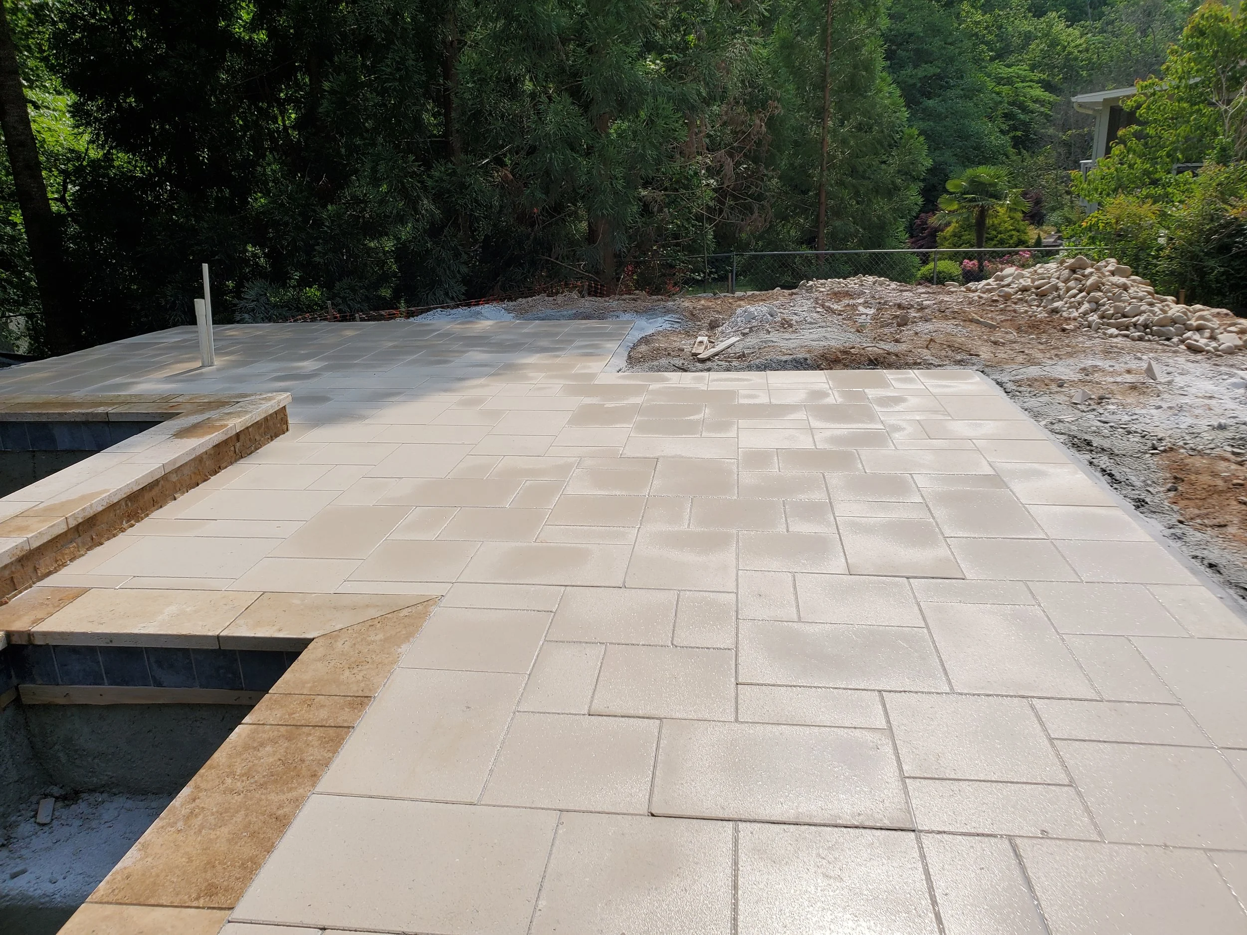 Construction site with a partially paved outdoor area, dirt, rocks, and surrounding greenery.