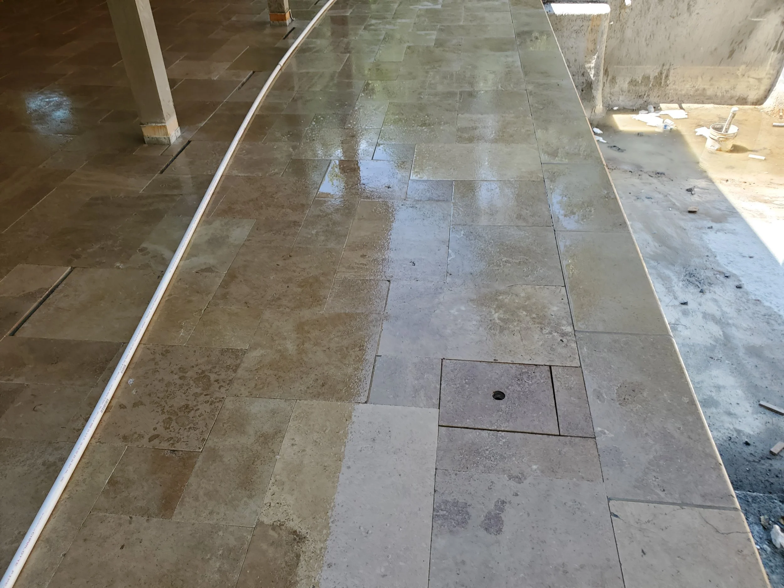Wet floor tiles in the process of being cleaned or installed, with a white pipe running across the tiles and some construction tools and debris visible in the background.