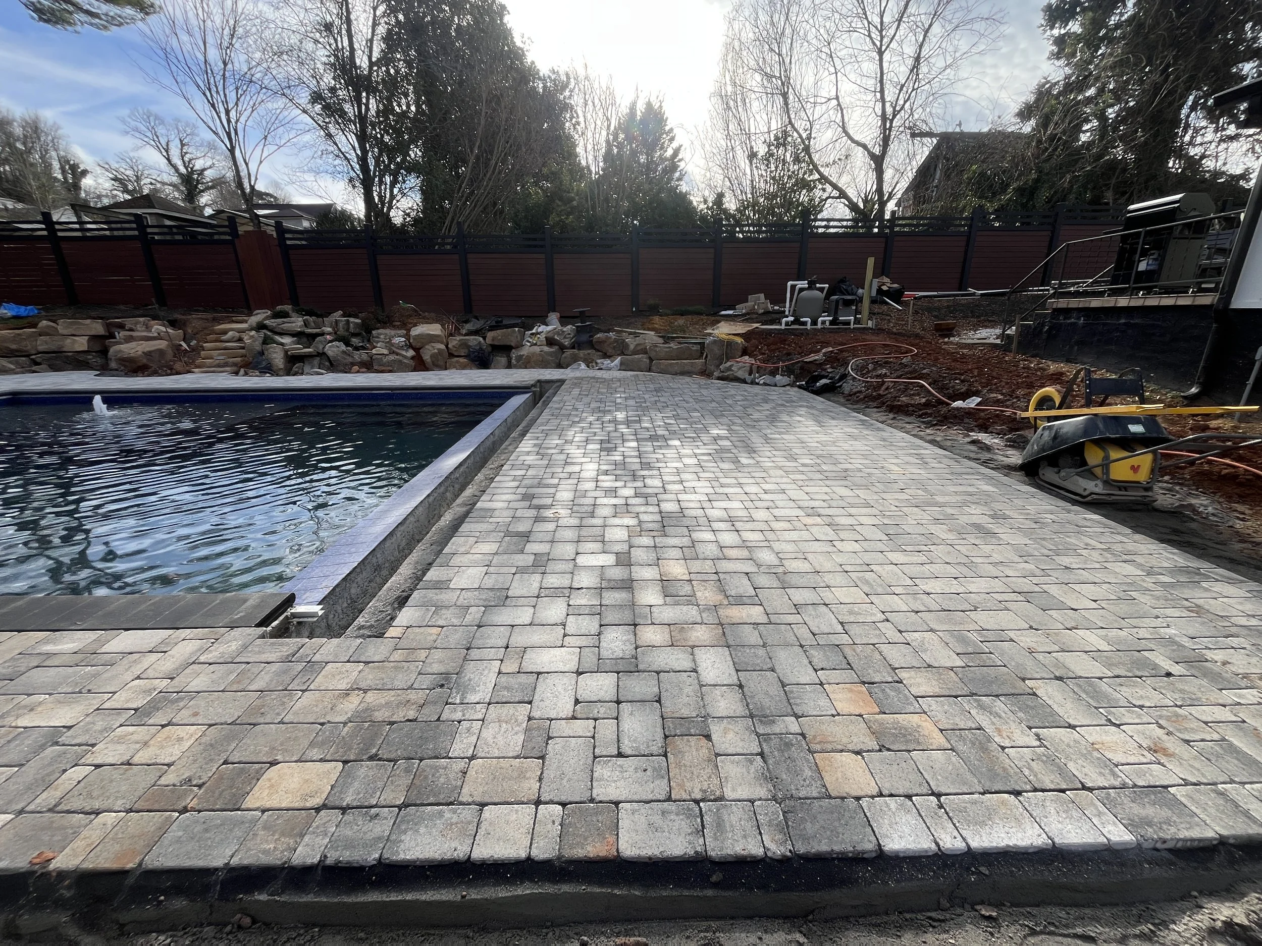 Backyard landscape under construction with a paved patio, a swimming pool, a stone retaining wall, and a wooden fence, with construction tools and materials scattered around.