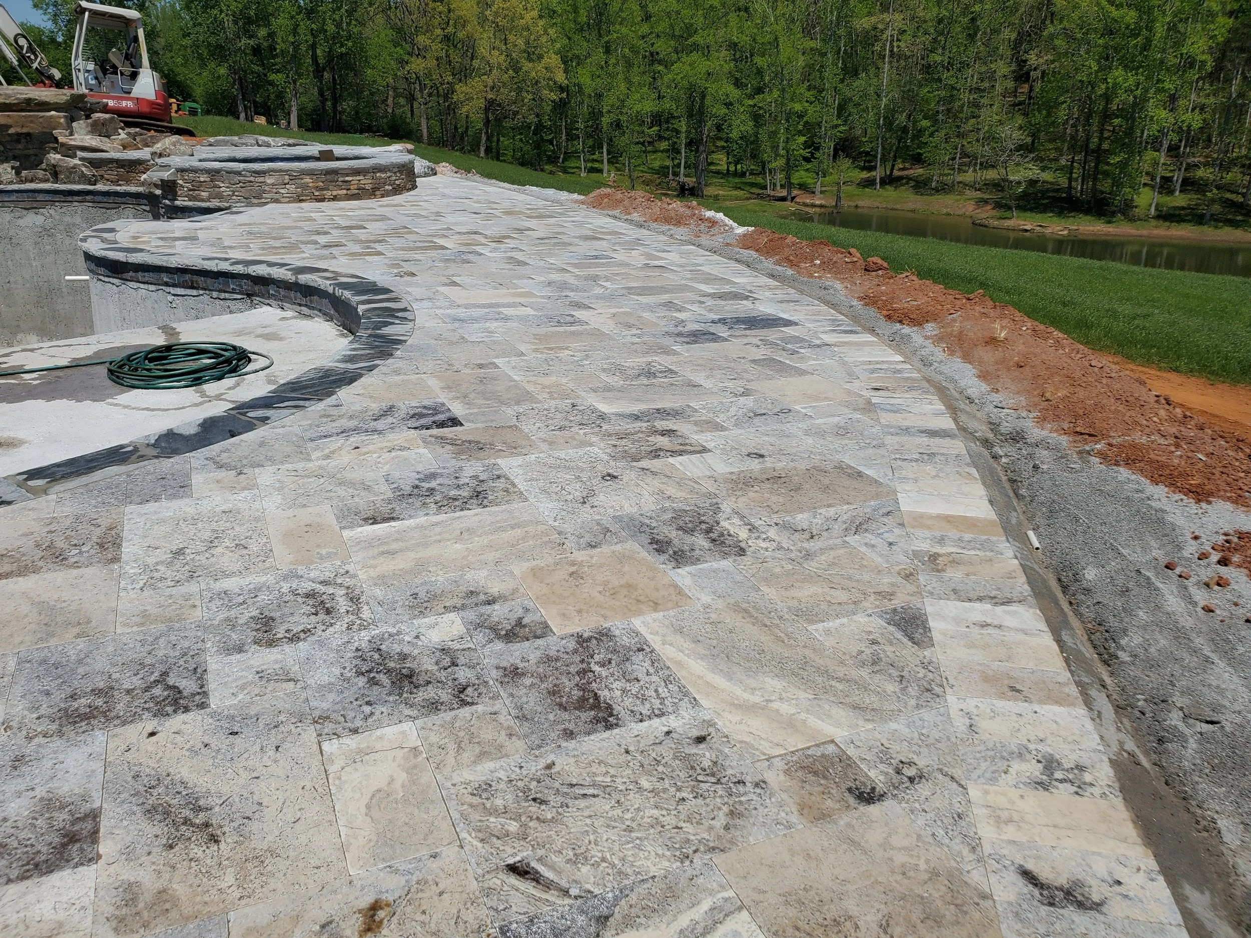 A newly constructed outdoor stone patio with a curved design, set beside a grassy area and a small body of water, with a portable excavator and construction materials in the background.