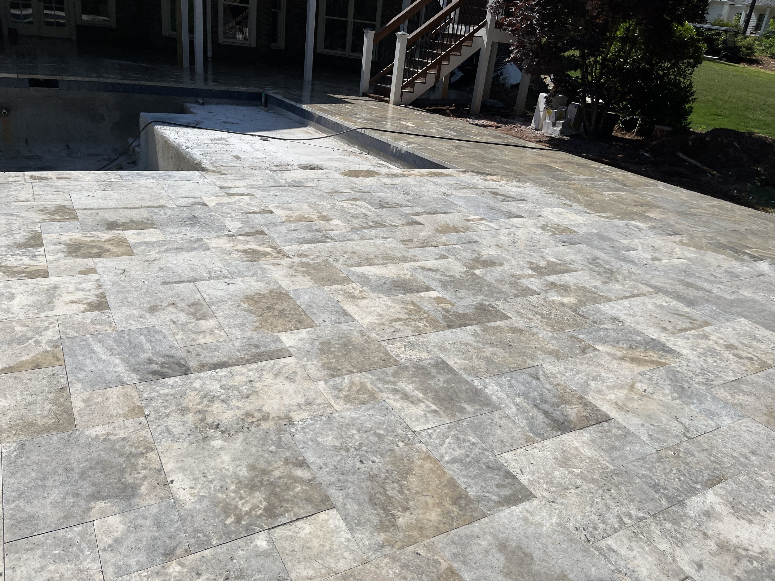 An outdoor backyard patio with stone tiles, a staircase leading to a deck, and a partially empty swimming pool with construction debris around.