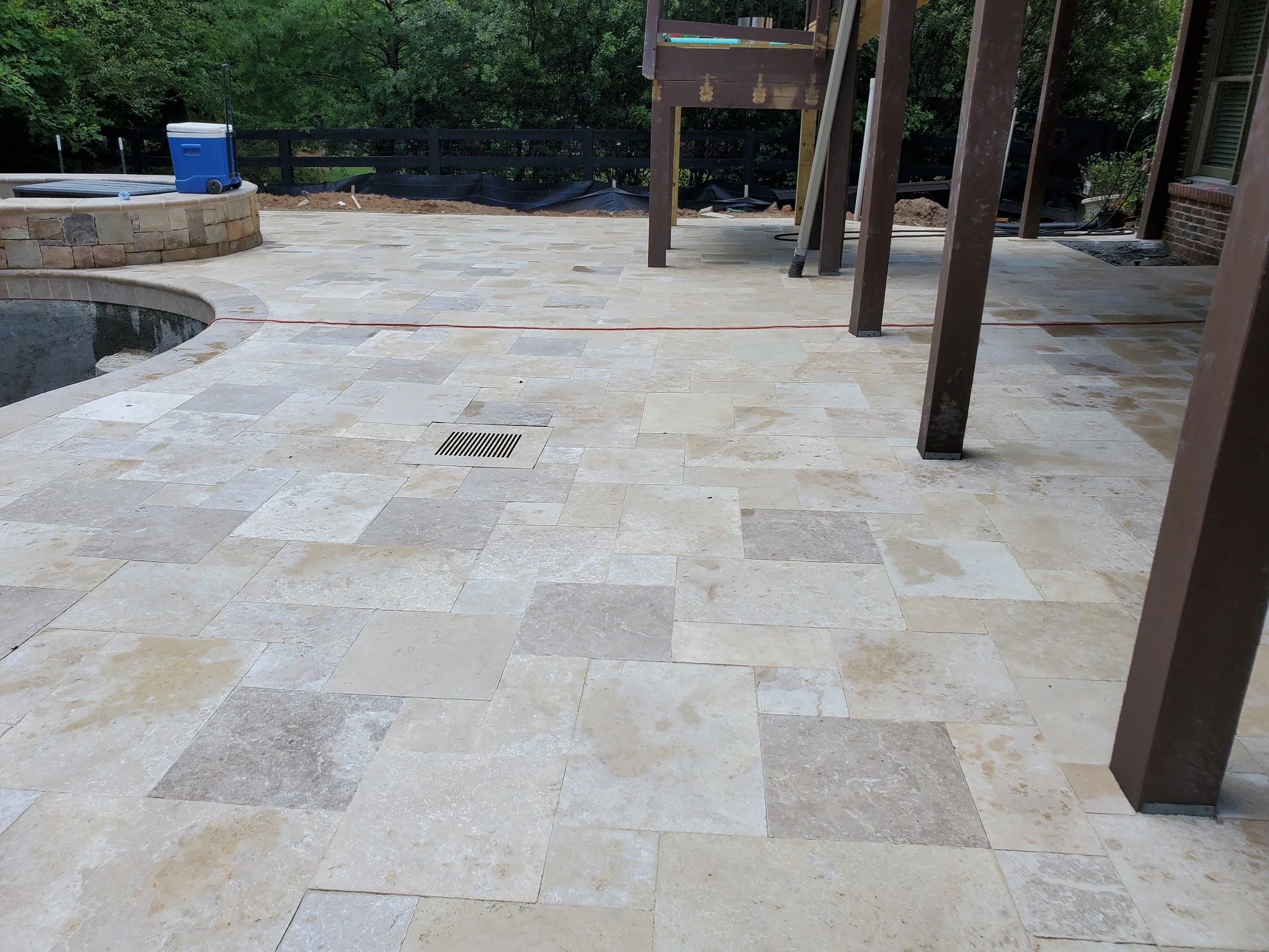 A backyard patio under construction with beige pavers, a raised stone fire pit, and a wooden deck with a support post, surrounded by trees and greenery.