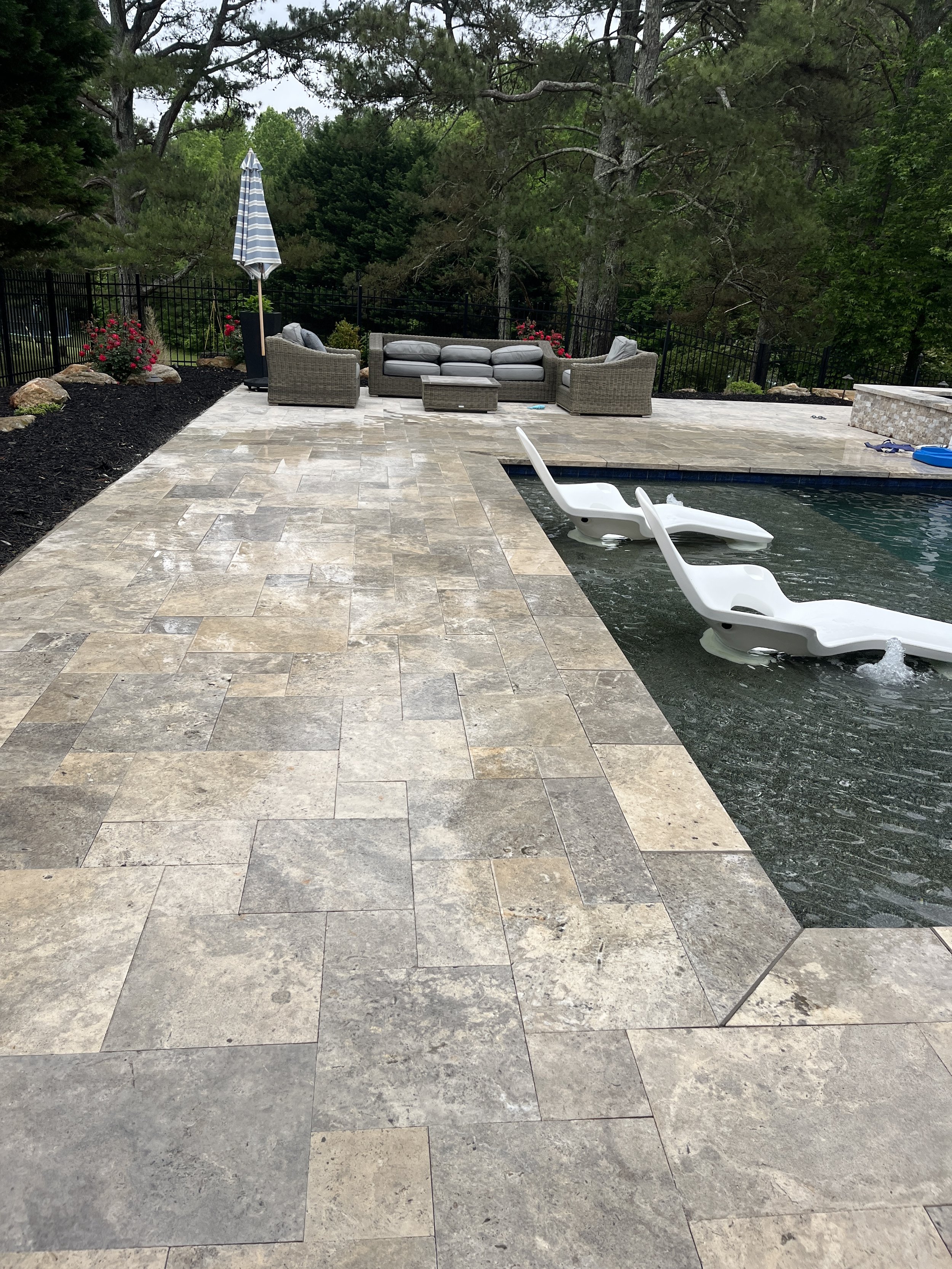 A backyard patio with stone tiles, a swimming pool with a white lounge chair in the water, outdoor sofas with pillows, a patio umbrella, and surrounding greenery including trees and flowering plants.