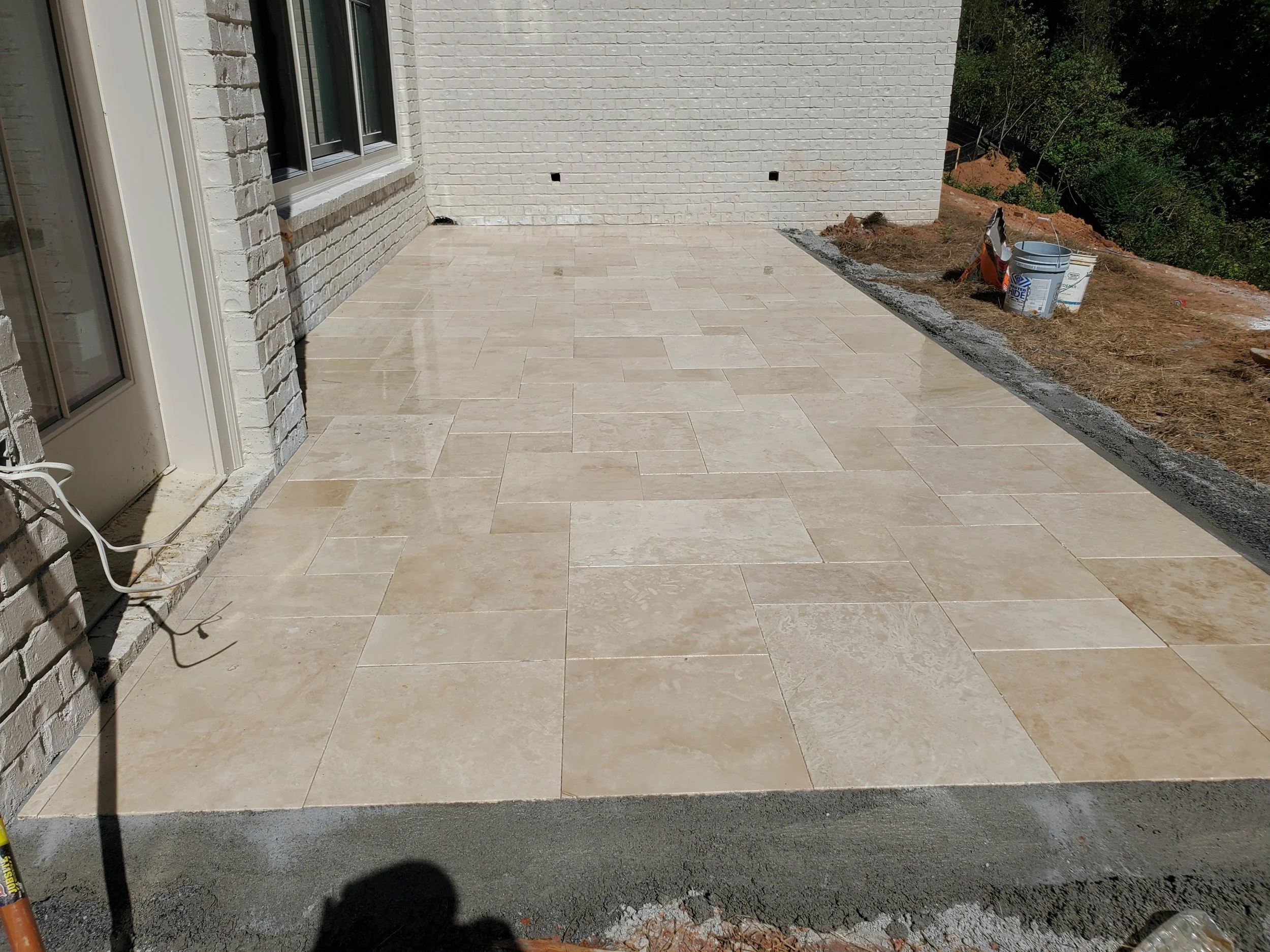 Newly constructed patio with beige tiles adjacent to a brick house, with construction tools and materials on the side, and a wooded area in the background.