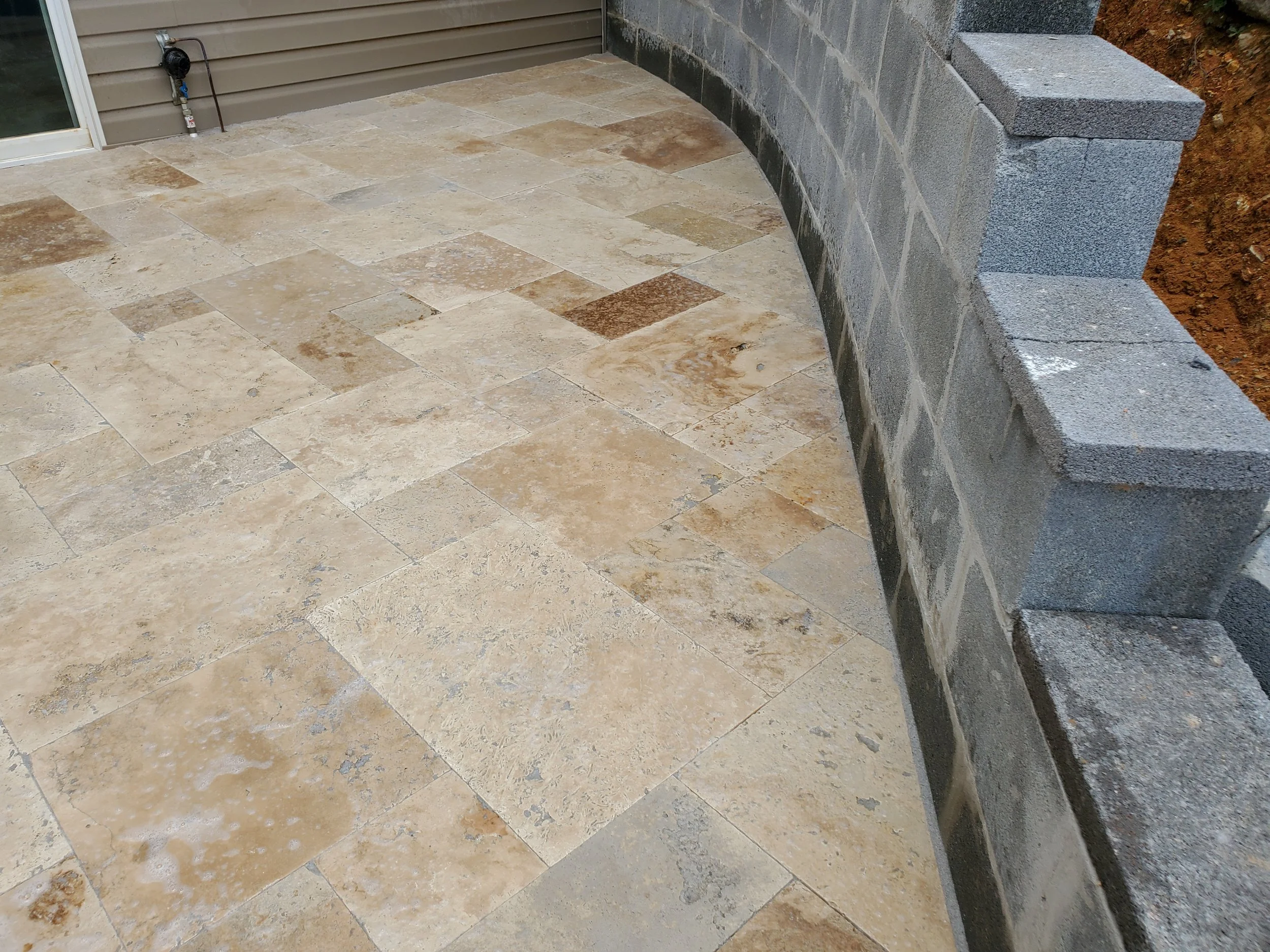 A newly constructed outdoor patio with beige and brown stone tiles and a gray cinder block wall, next to a house with beige siding and a sliding glass door.