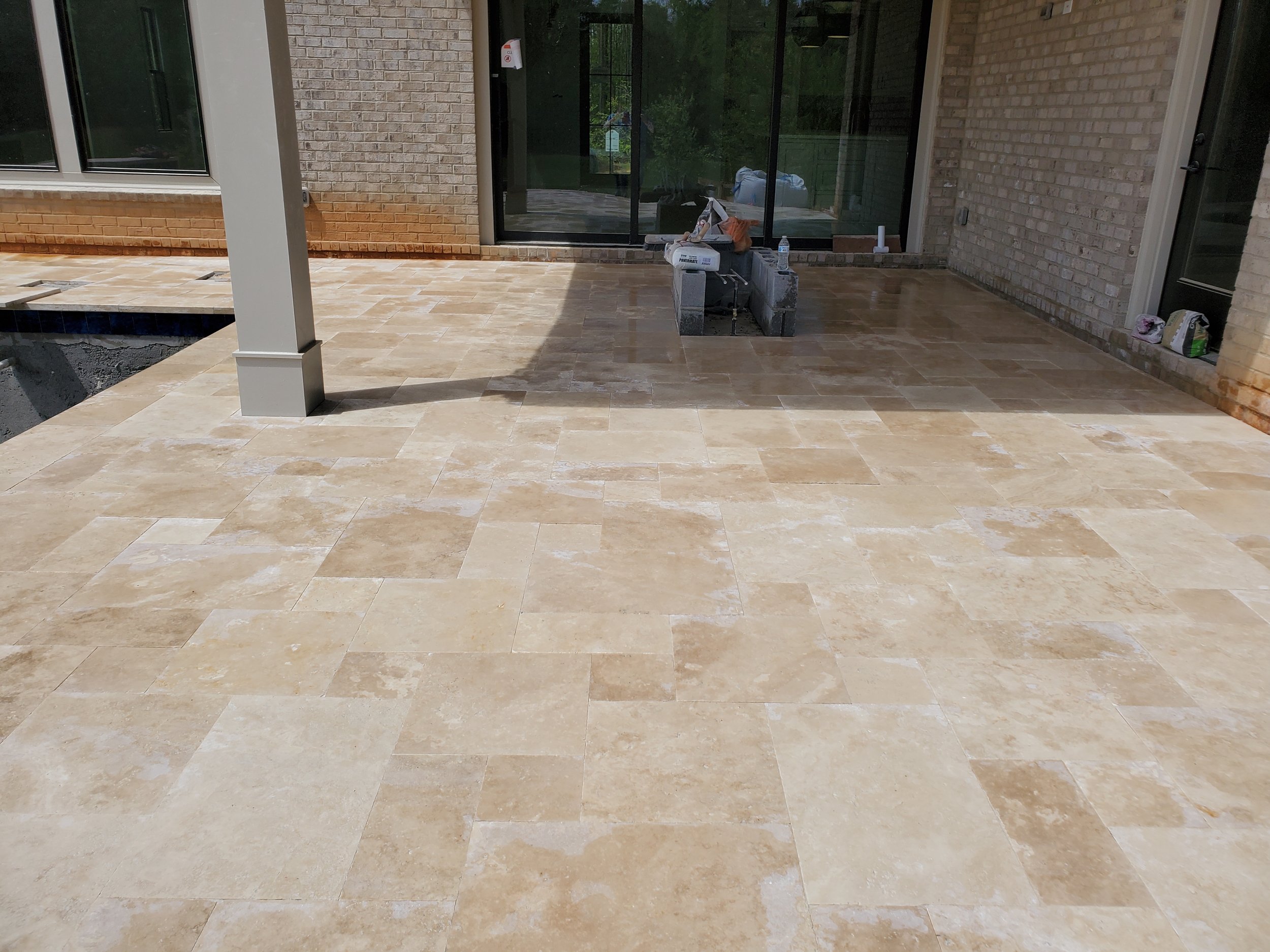 Back patio or porch area with beige stone tiles, brick walls, large glass sliding doors, construction supplies, and partially completed features.