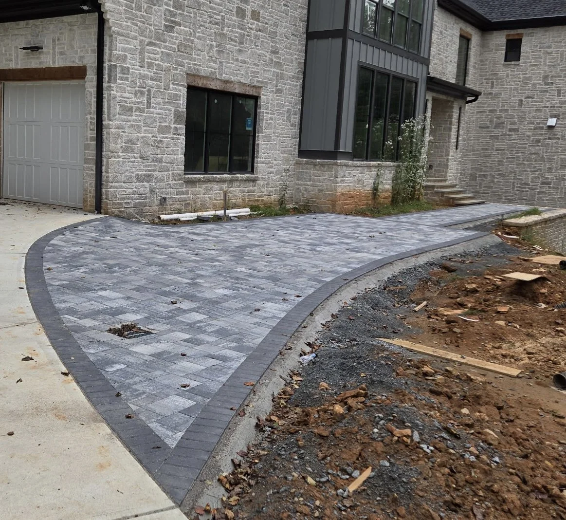 Newly paved gray brick driveway curving towards a house with stone walls and steps, surrounded by dirt and construction debris.